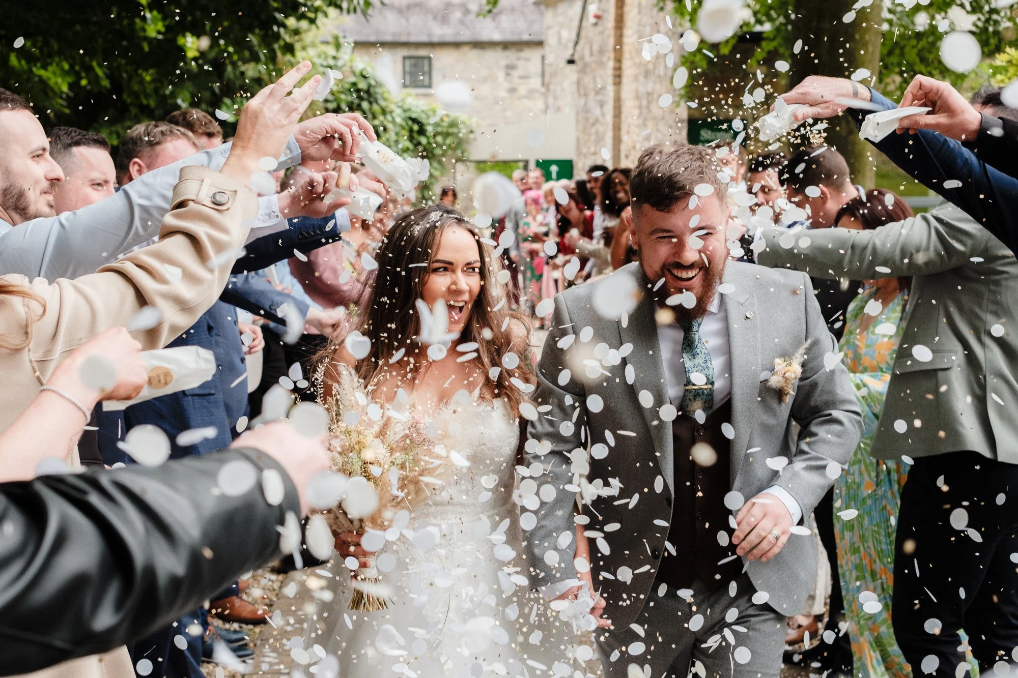 Bride and groom walking through confetti as guests throw confetti at their wedding celebration outdoors.