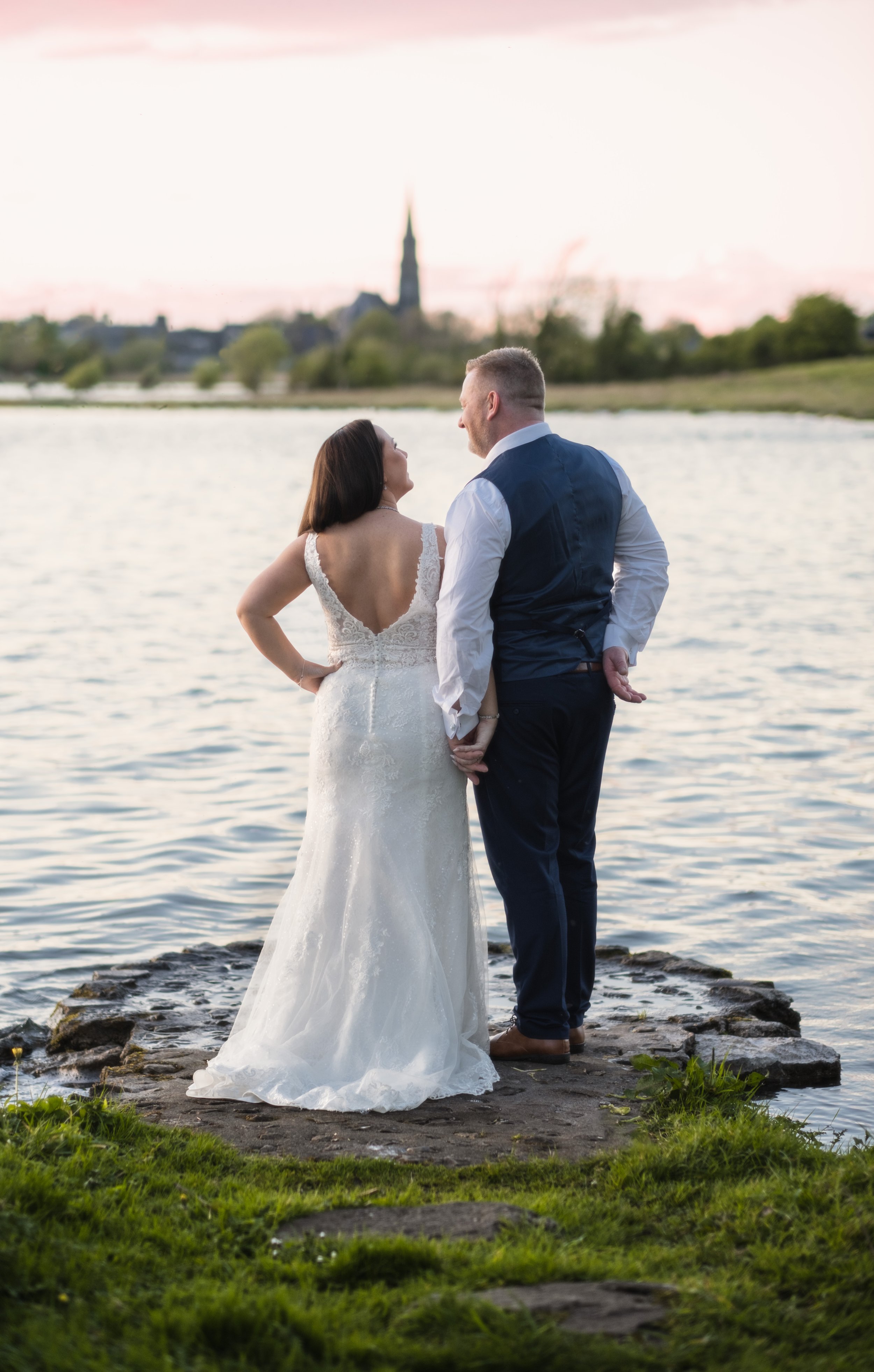A bride and groom standing on rocks by the water at sunset, holding hands and looking at each other, with a city skyline and church steeple in the background.