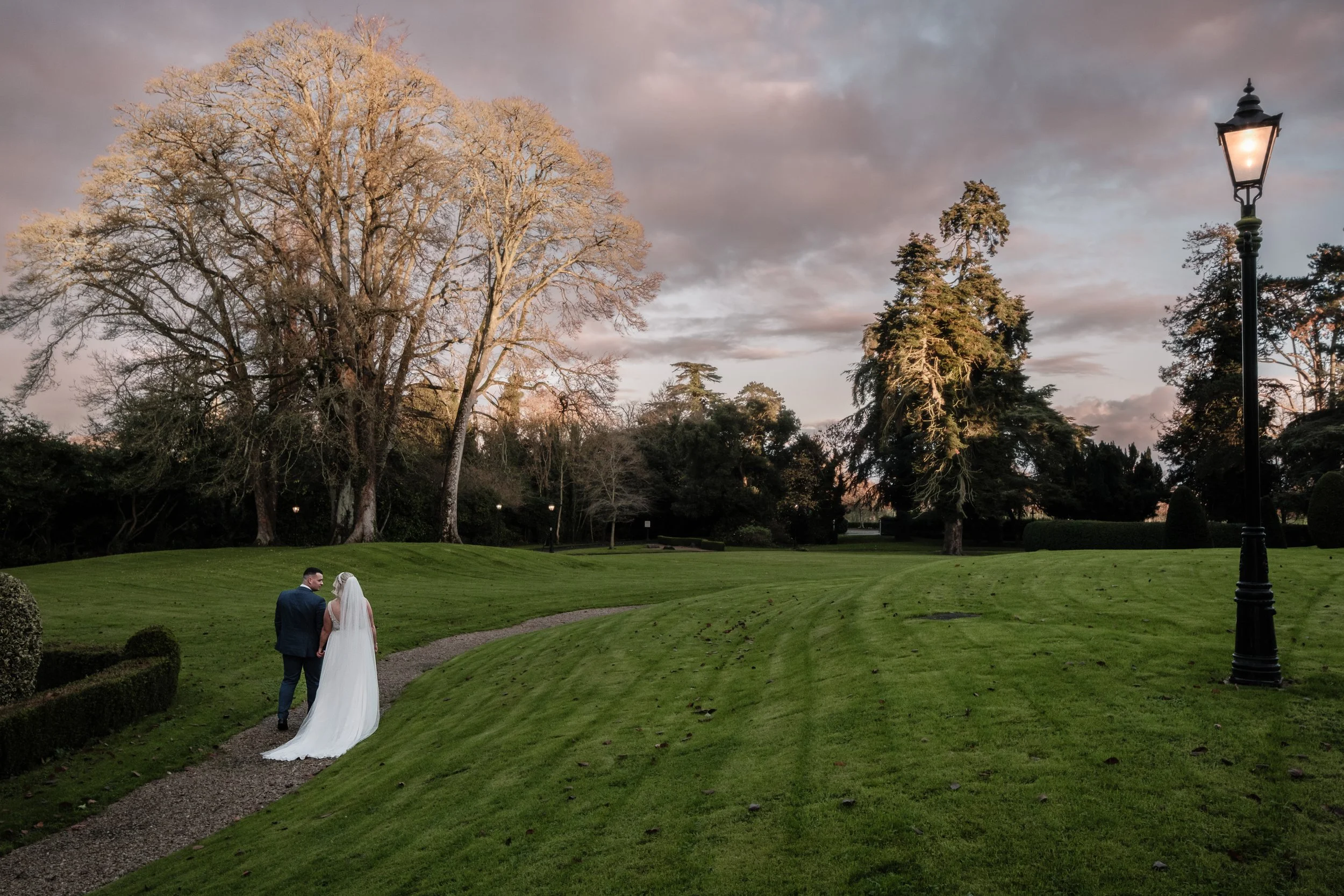 Bride and groom walking on a path in a park with green grass, trees, and a lamppost under a cloudy sky.