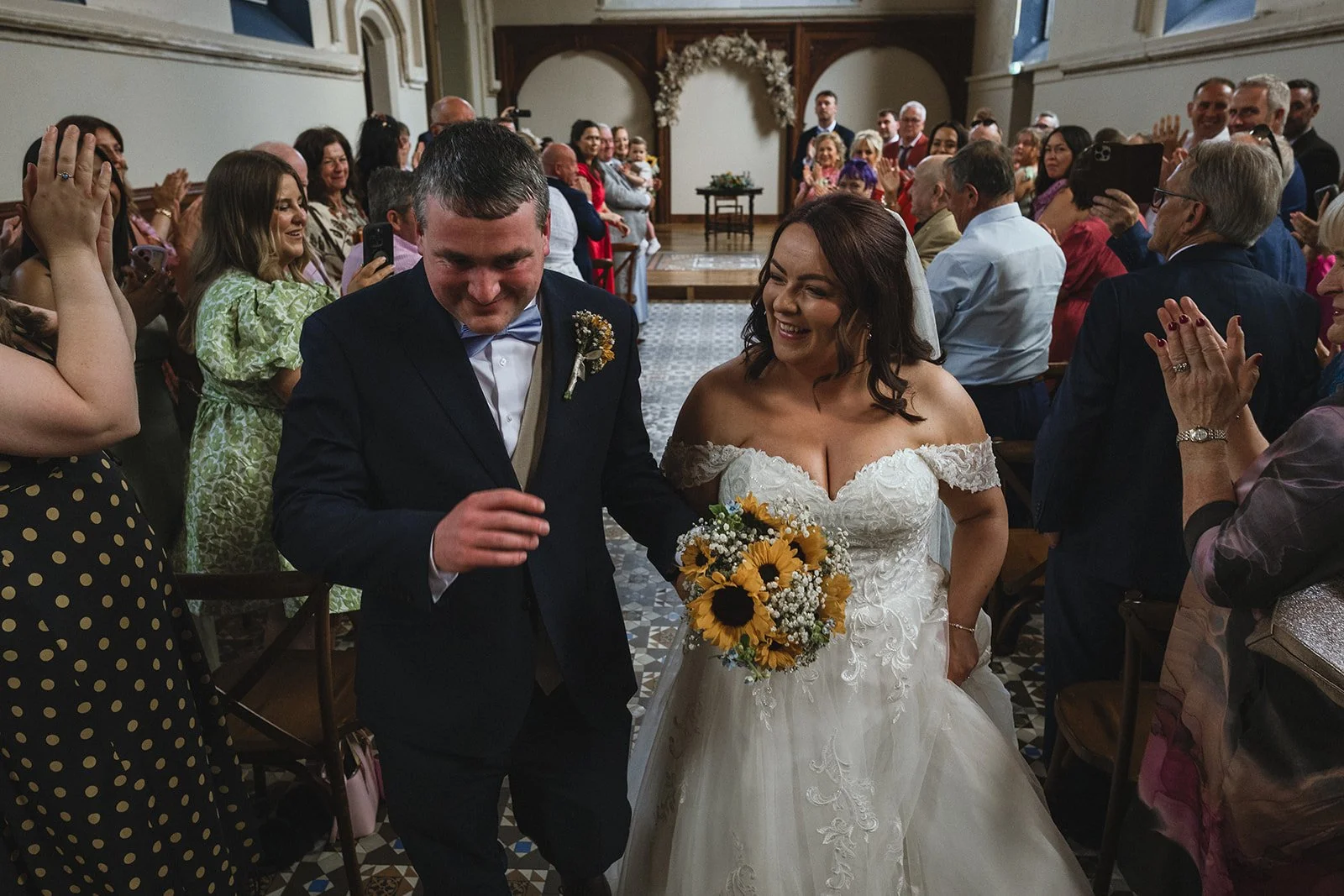A bride and groom smiling and walking down the aisle at a wedding ceremony. The bride is holding a sunflower bouquet, and the groom is dressed in a dark suit with a bowtie. Guests are sitting and standing on either side, capturing the moment with pho