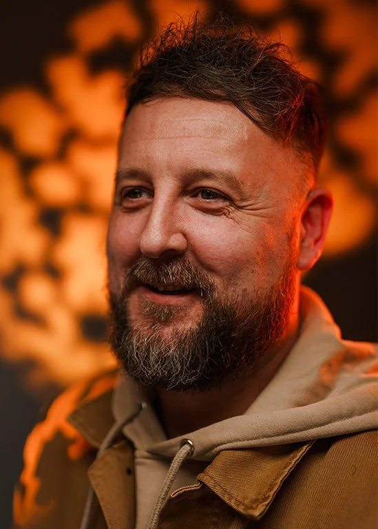 A smiling man with a beard and short hair, wearing a beige hoodie and brown jacket, standing in front of a fiery, orange background.