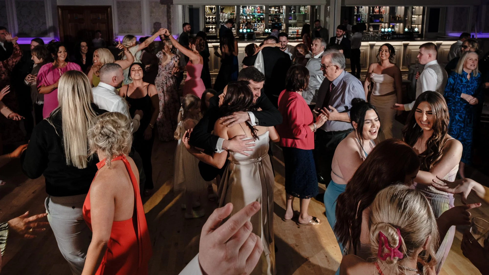 People dancing and celebrating at a lively party or wedding reception in an elegant venue with a bar in the background.