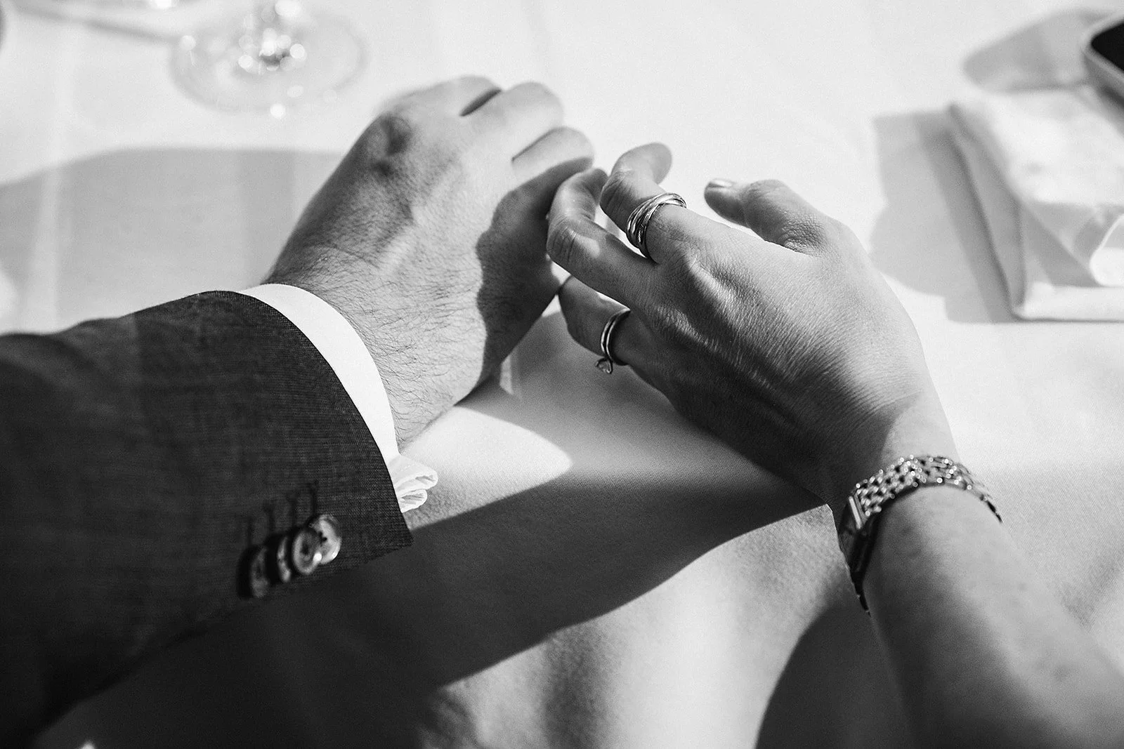 Close-up of a man and woman holding hands, with the man's hand resting on top of the woman's hand, on a table. The man is wearing a dark suit, and the woman has jewelry on her wrist and rings on her fingers.