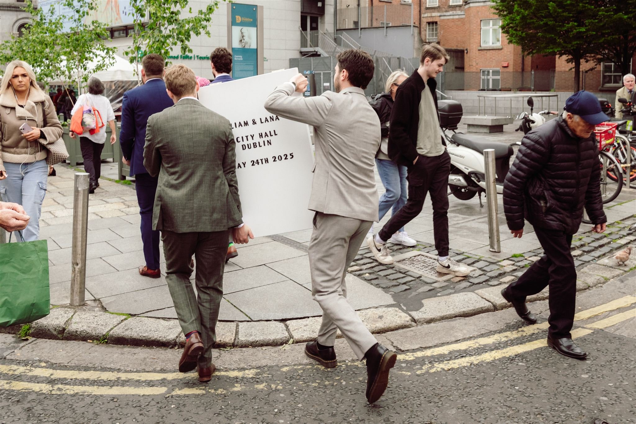 People walking on a city sidewalk, some carrying signs with text about Dublin city hall event on May 24th, 2025. The scene includes individuals dressed in casual and business attire, parked motorcycles, and bicycles in the background near some buildi