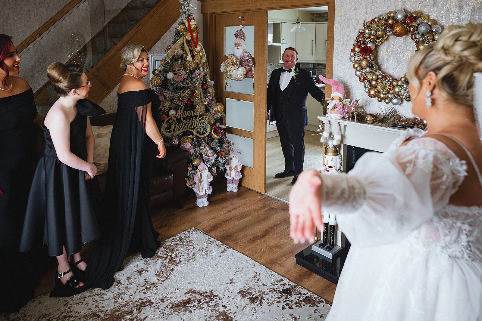 A bride in a white wedding dress extending her arm toward a man in a tuxedo, with three women in black dresses, all smiling, standing near a decorated Christmas tree and holiday decorations.