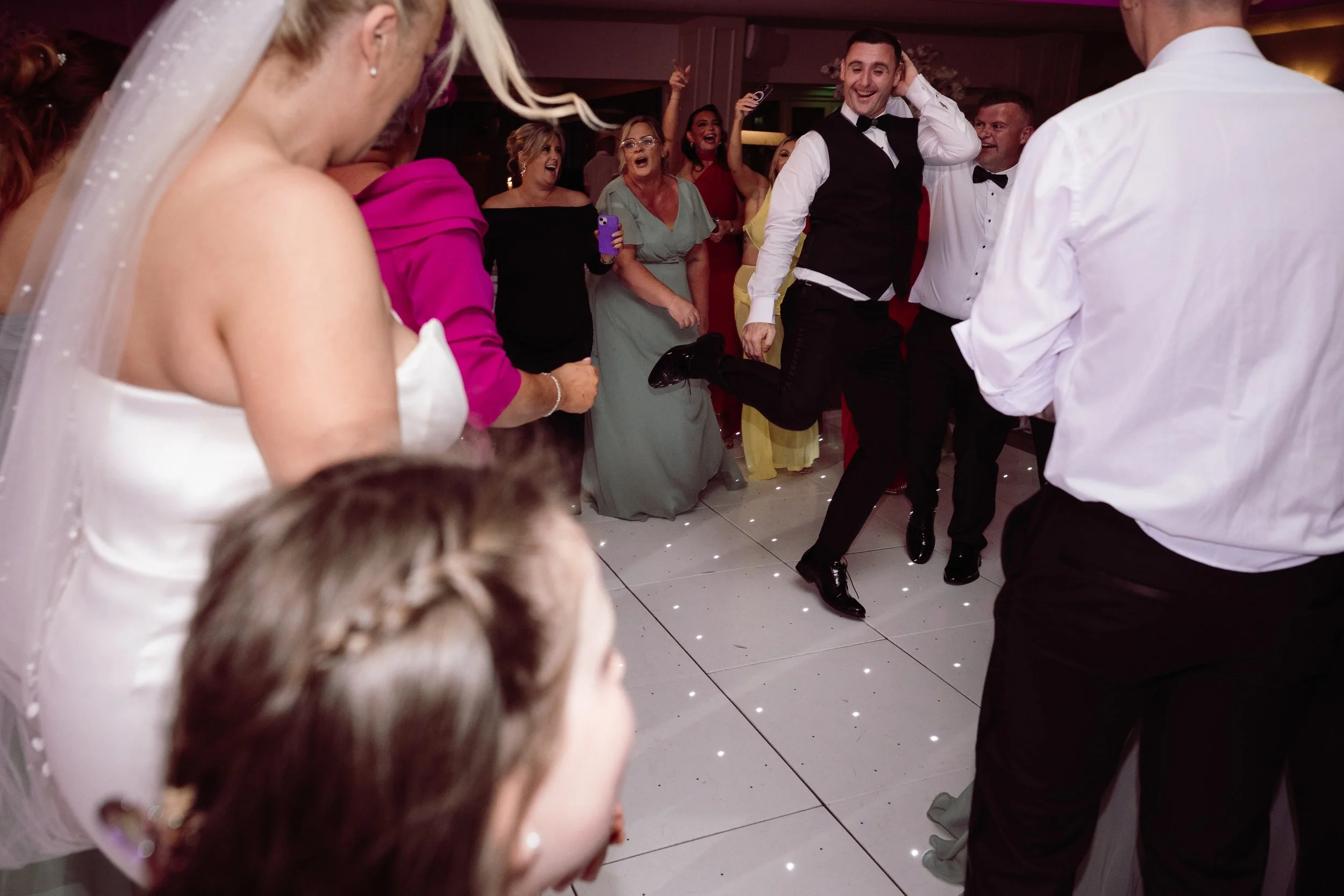 People dancing and enjoying at a wedding reception, with the groom prominently smiling and kicking up his leg.