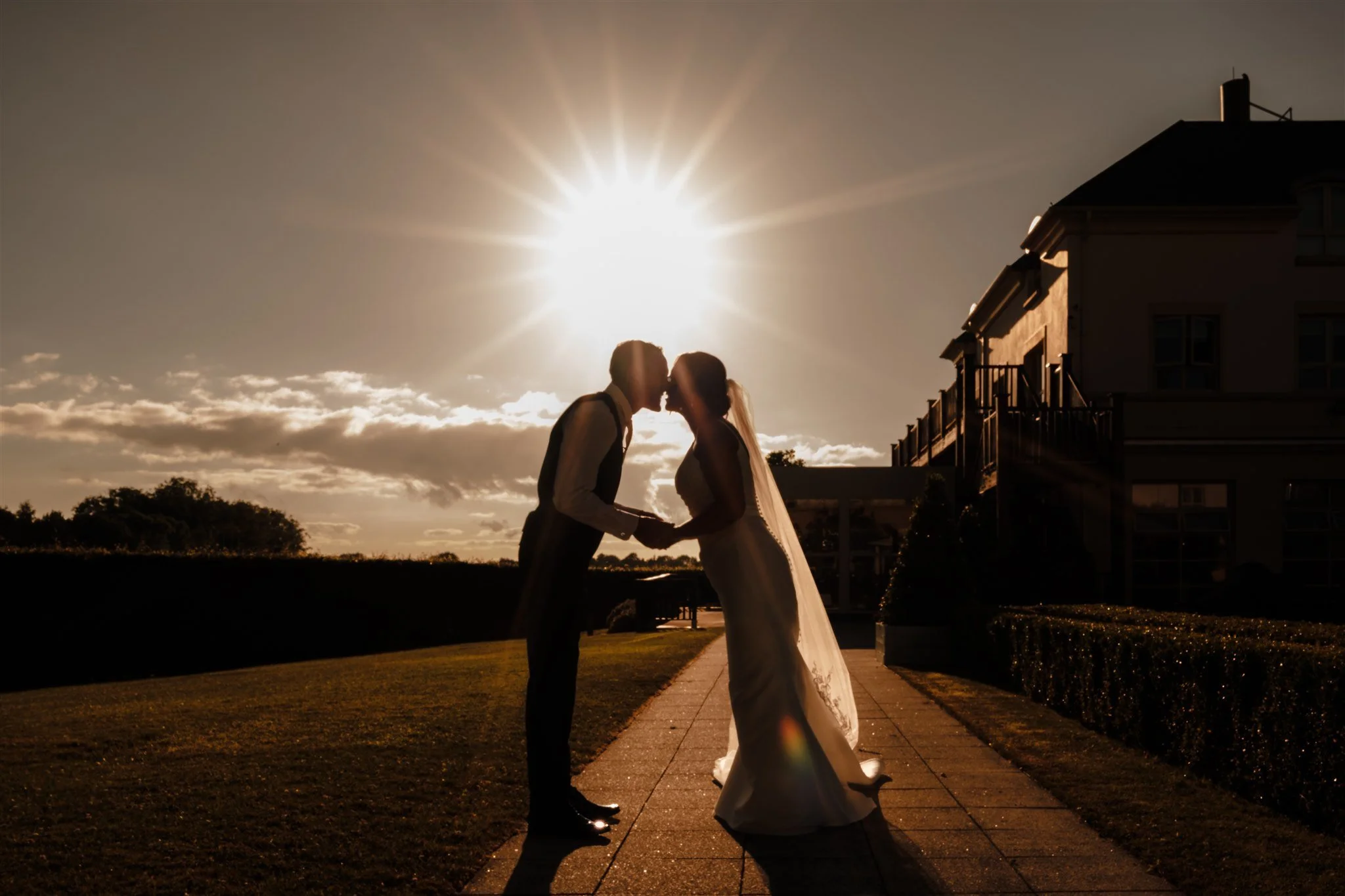 A bride and groom sharing a kiss during sunset, with the sun creating a silhouette behind them. The scene takes place outside near a building with multiple floors.