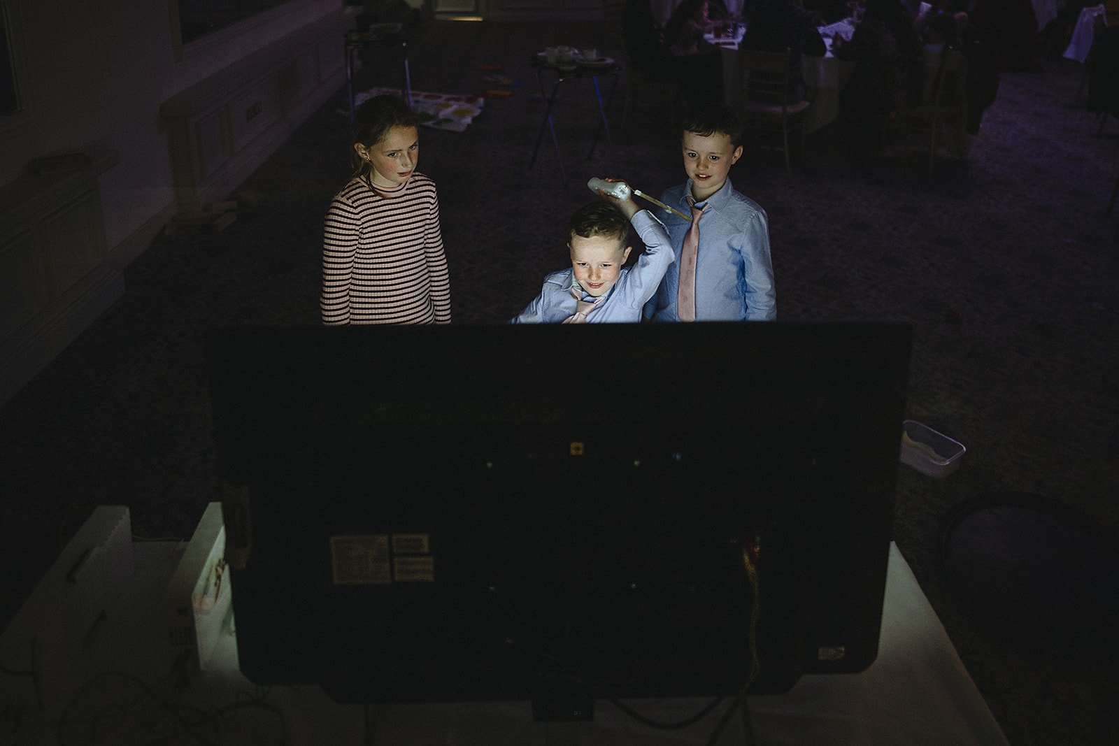 Three children, two boys in shirts and ties and one girl in a striped shirt, watching a large screen in a dark room, with one boy holding a remote control.