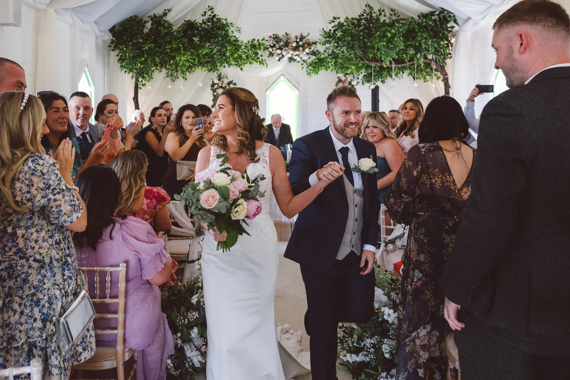 Bride and groom exchanging vows at a wedding ceremony inside a decorated venue with guests watching and taking photos.