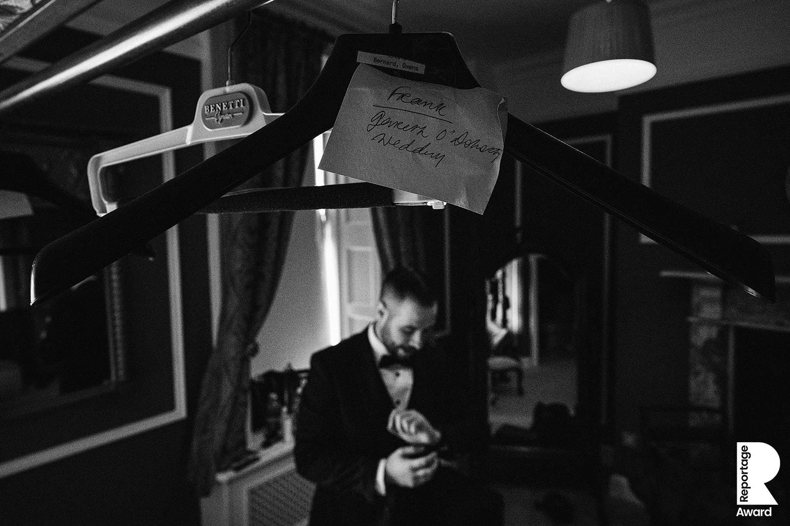 Black and white photograph of a man in a tuxedo in a dimly lit room, seen through a clothing hanger with a note attached. The note reads 'Frank Gowers O'Donnell Wedding'.