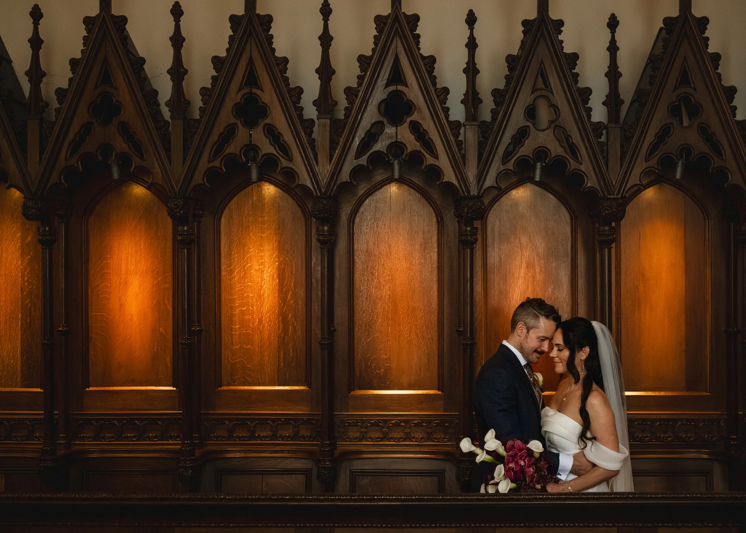 A couple in wedding attire sharing a close moment in front of a dark wood Gothic-style wall with pointed arches and intricate details.