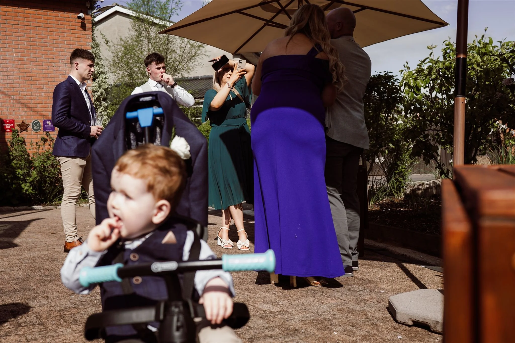 Group of people at an outdoor celebration, including two women and a man in formal attire, a young boy sitting in a stroller, and another young man with a drink, under a large umbrella, with a brick building and greenery in the background.