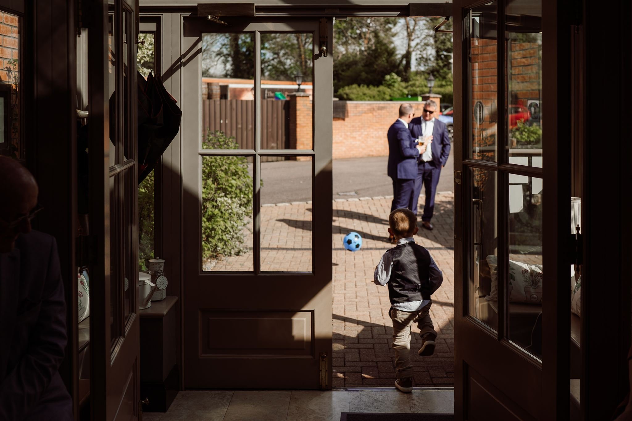 A young boy running outside through a door towards two men in suits, who are standing and talking in a yard with a brick wall, trees, and parked cars. A blue soccer ball is on the ground near the boy.