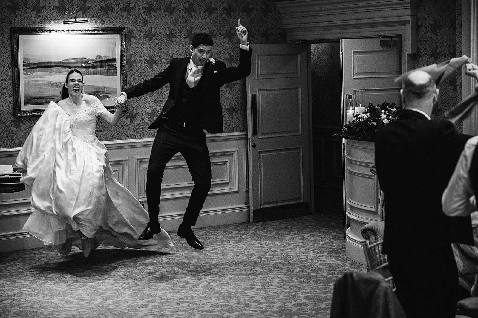 Black and white photo of a bride and groom dancing and jumping on the floor in a room decorated for a wedding reception.