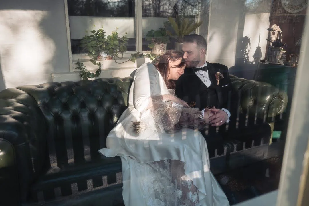 A couple dressed in wedding attire sitting on a dark green leather couch, leaning on each other inside a sunlit room with potted plants and decorative items in the background.