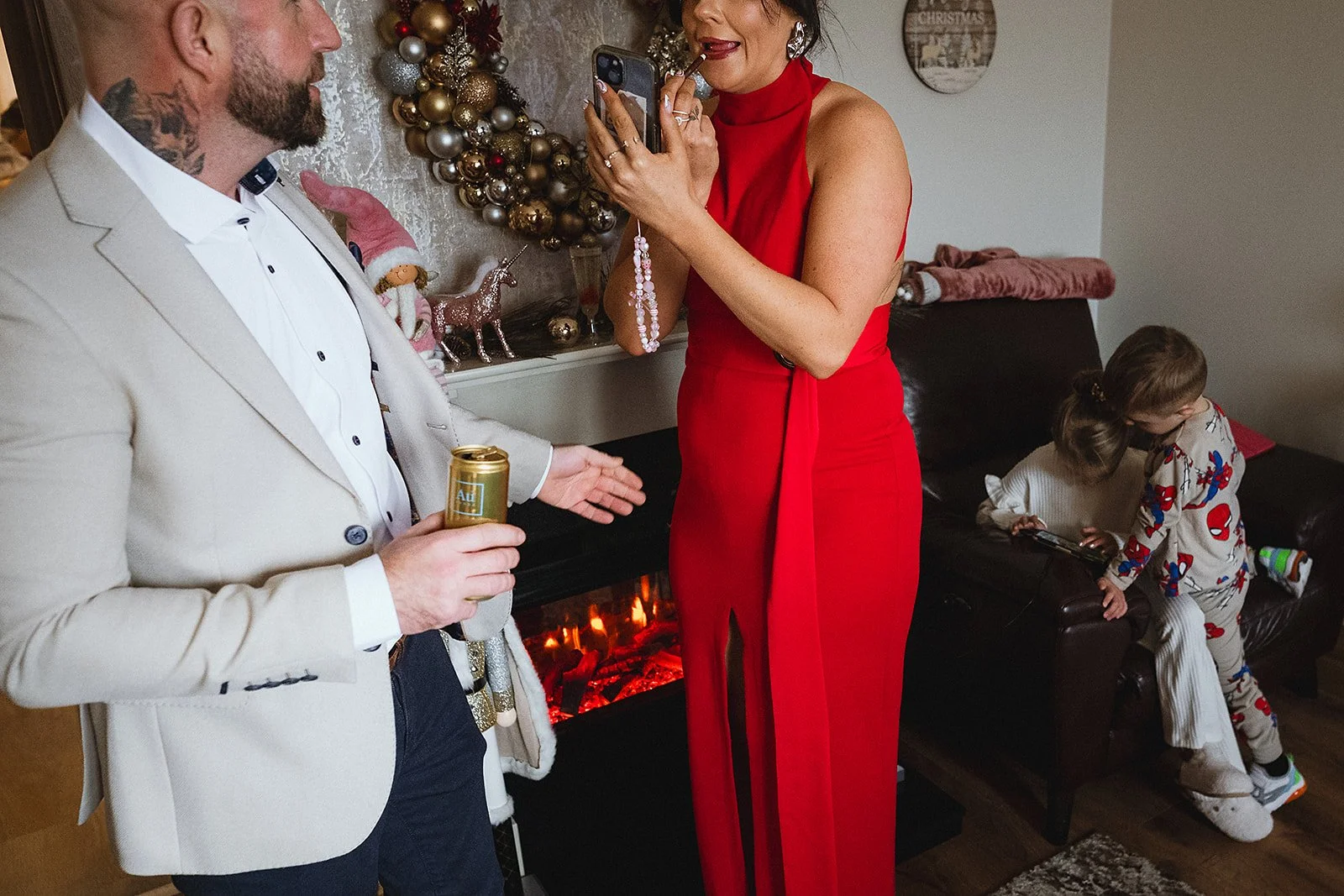 A man in a beige suit with a tattoo on his neck and a white shirt, holding a can, is talking to a woman in a red dress, who is smoking and holding a phone. Two children in pajamas are on a brown couch, with one playing with a toy. Decorative Christma