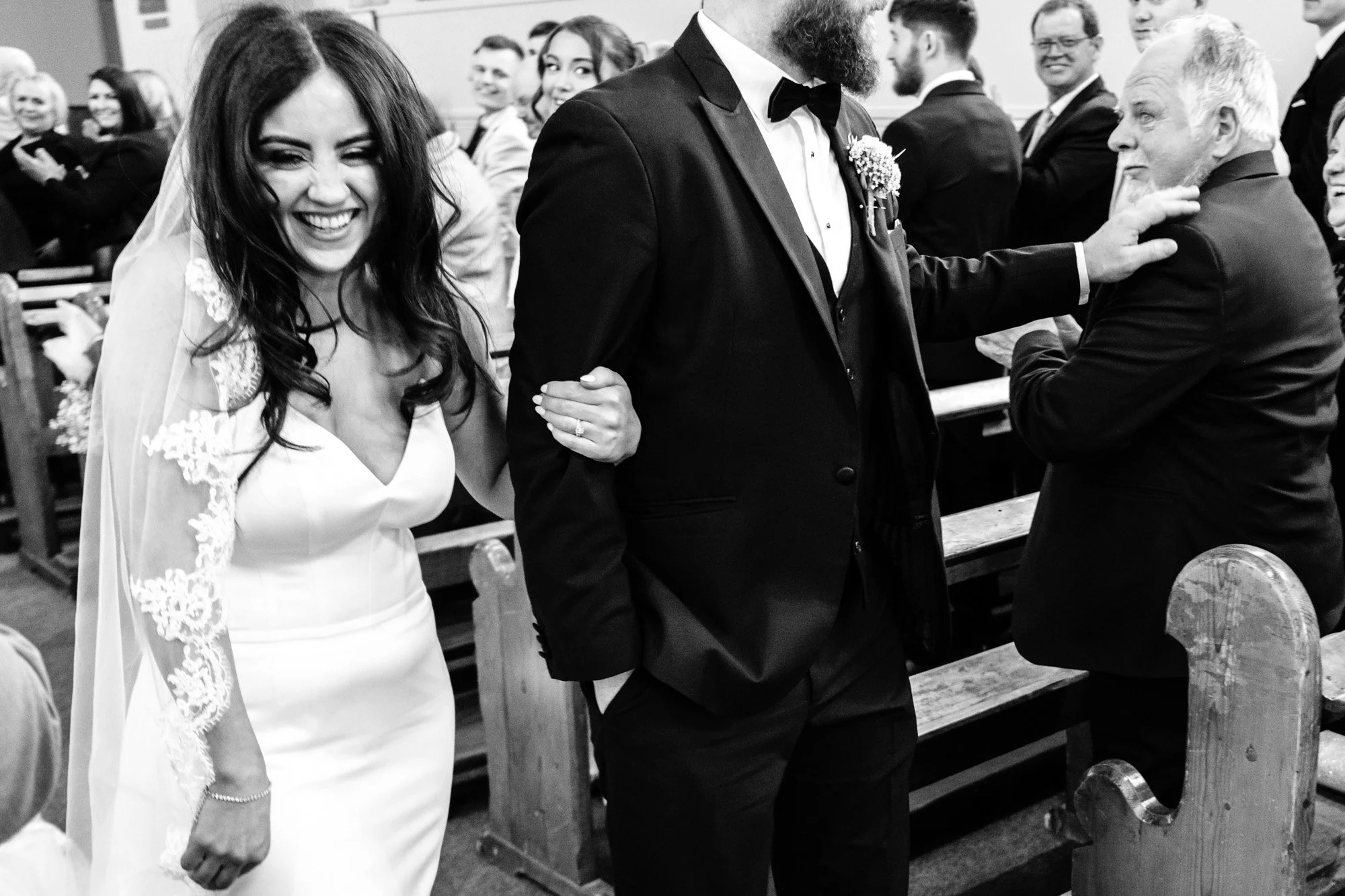 A black-and-white photo of a wedding ceremony showing a smiling bride with dark hair in a lace-embellished wedding dress, holding hands with a groom in a tuxedo. Both are standing in a church filled with seated guests, some smiling and others engaged