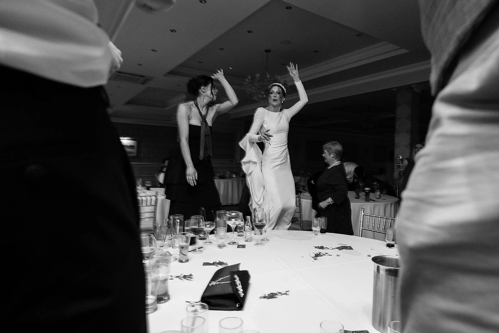 People celebrating at a wedding reception, with two women dancing on the table. One woman in a white dress is dancing with her hands in the air, and another woman in dark clothing is nearby. The table is decorated and has glasses and utensils.