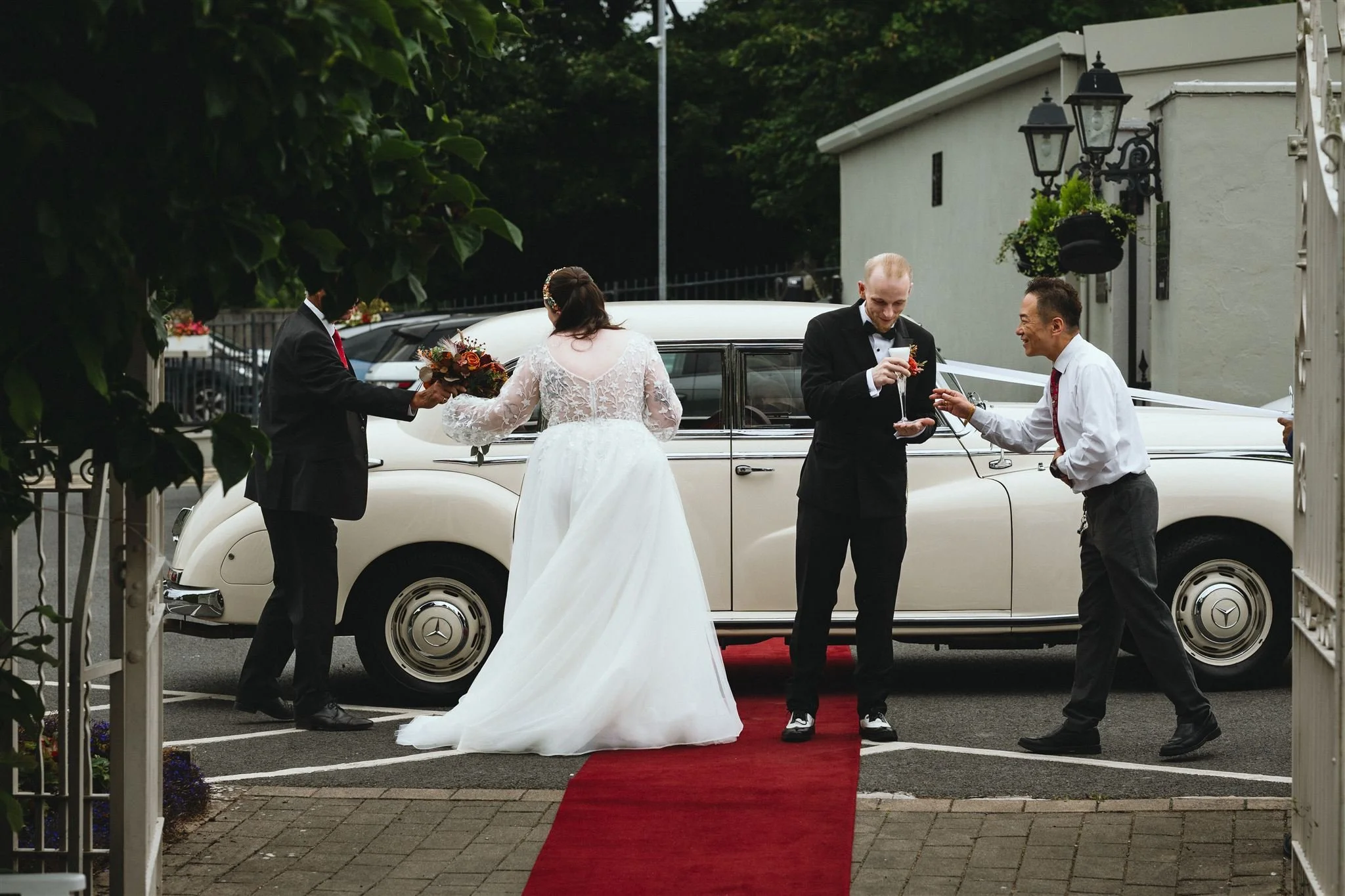A bride in a white wedding dress and three men, one of whom is the groom in a black suit, sharing a toast and exchanging gifts in front of a vintage cream-colored Mercedes-Benz car.