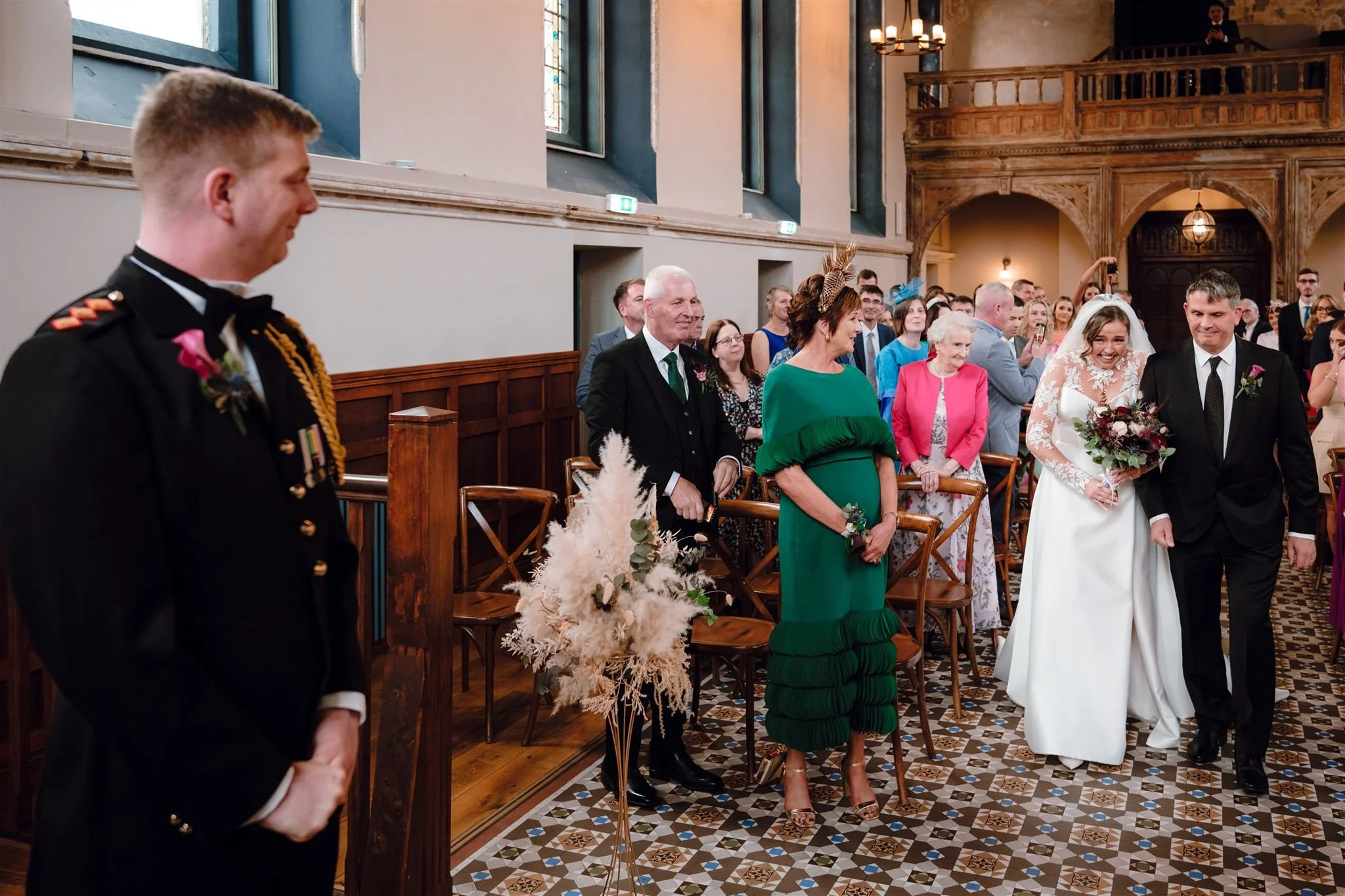 A bride and groom walk down the aisle in a church, smiling, with guests standing and watching. The bride holds a bouquet, and the groom has his arm linked with hers. The church has stained glass windows and wooden décor.