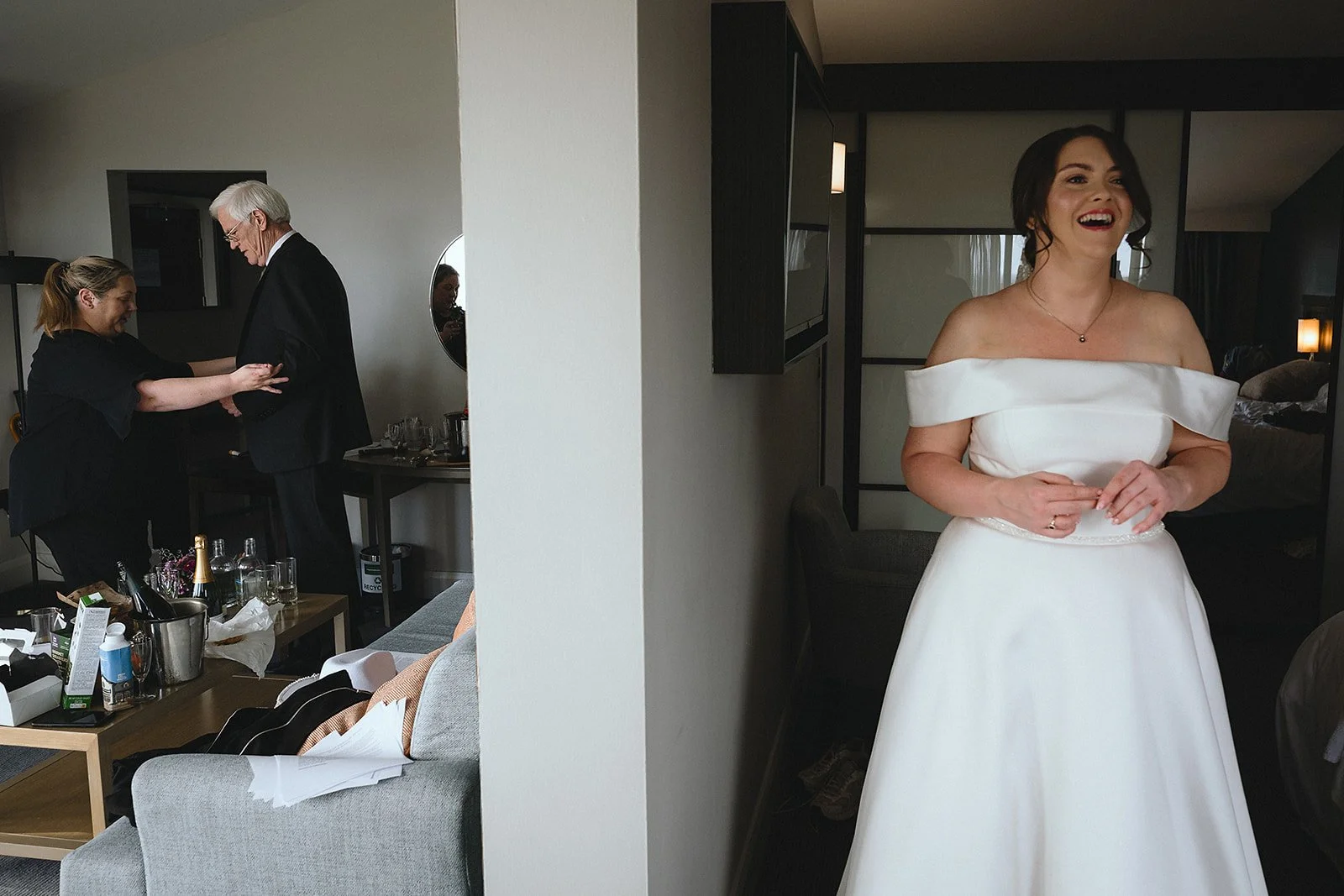 Partially divided hotel room with wedding preparations. On the left, a woman helps an older man in a suit. In the foreground, a sofa with a glass of water and papers. On the right, a smiling bride in a white off-shoulder wedding dress stands in front