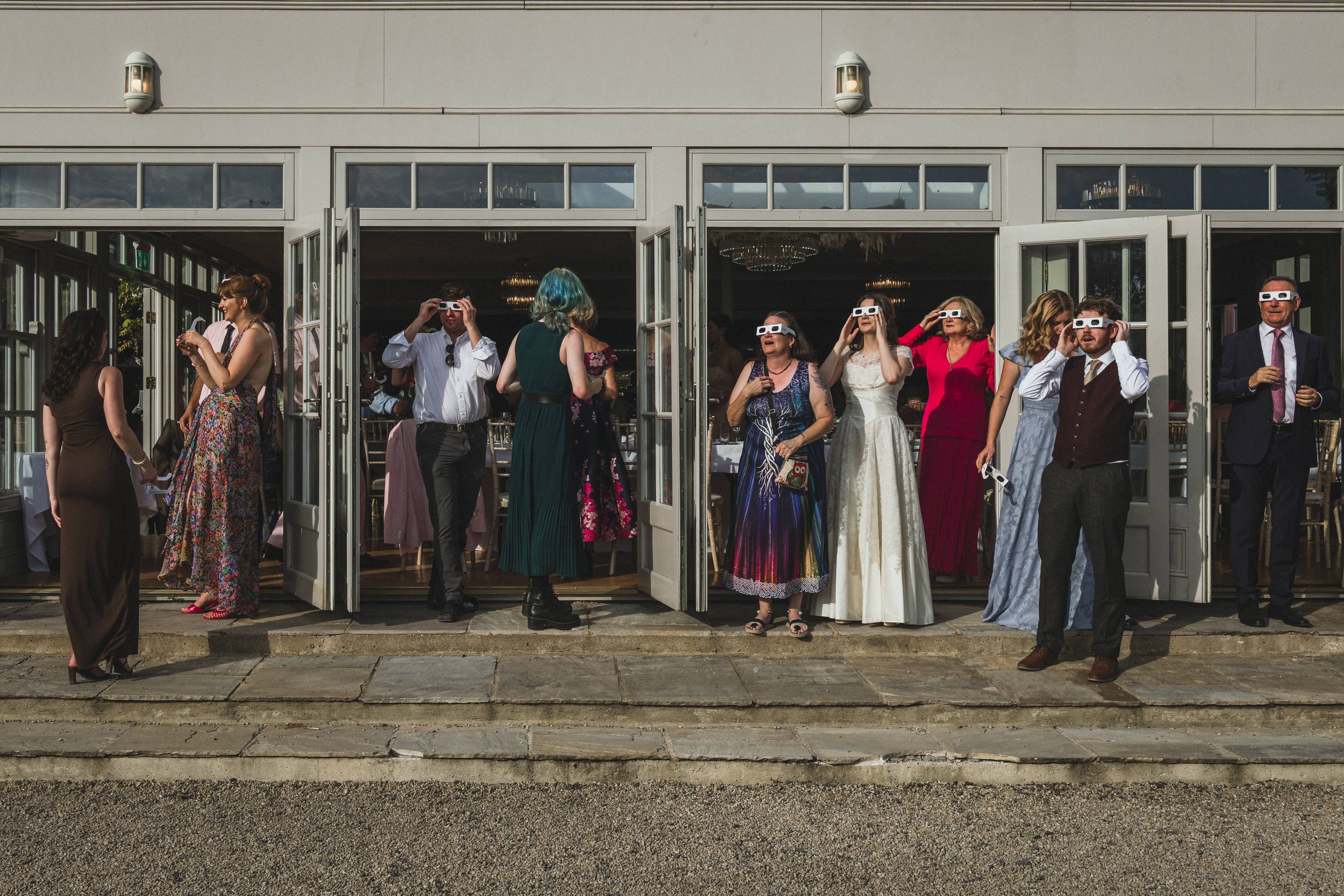 People gather outside a building, some wearing 3D glasses, likely at a wedding reception or celebration, with a mix of men and women dressed in formal and semi-formal attire.