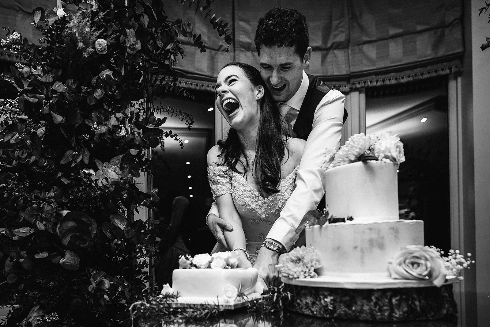 A newlywed couple cutting a wedding cake together, smiling and laughing, surrounded by floral decorations in a wedding reception setting.