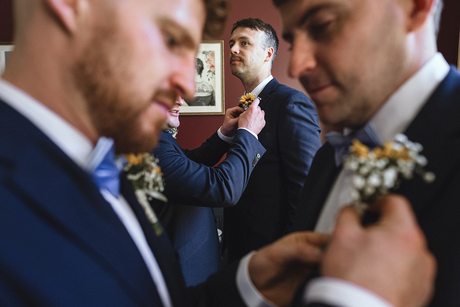 Four men dressed in tuxedos, with boutonnières, preparing for a wedding, focusing on the groom adjusting his boutonnière.