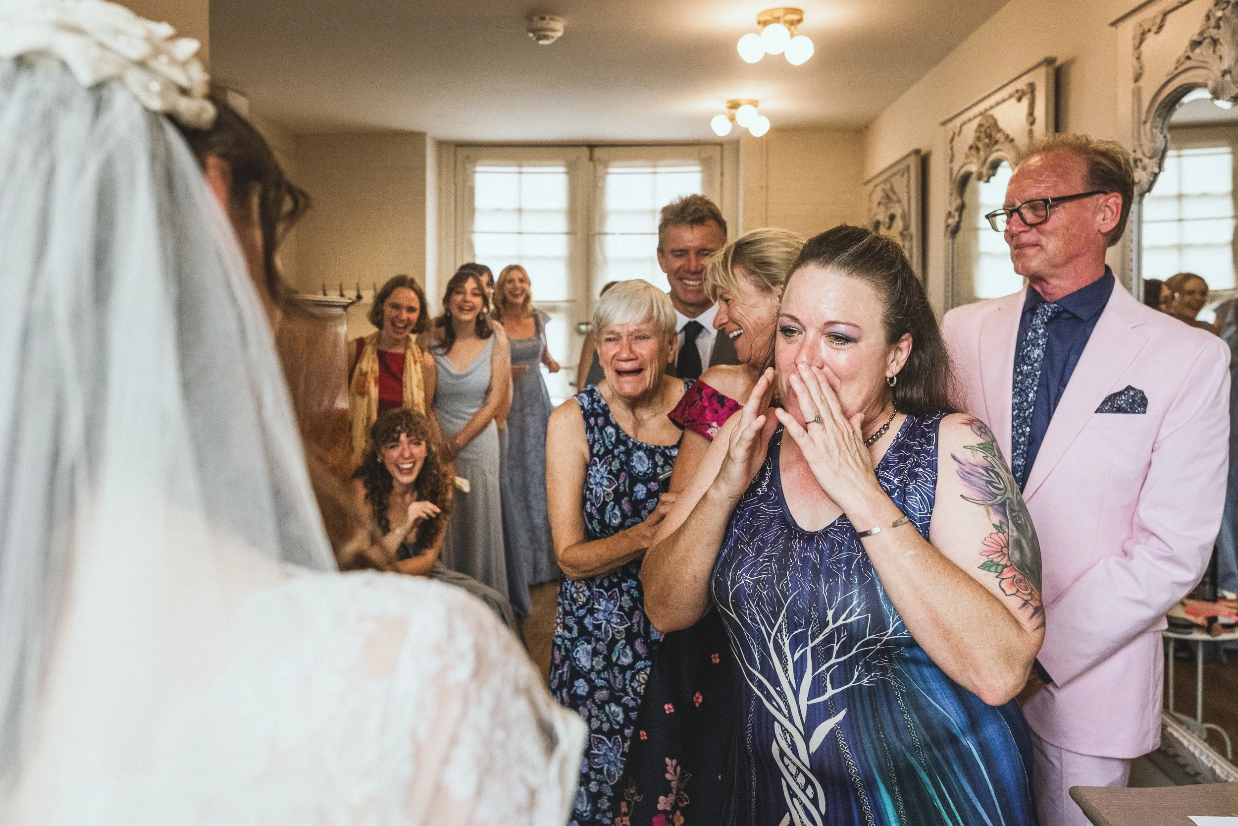 A group of people celebrating a wedding ceremony, with women crying tears of joy, and the bride's dress visible in the foreground.
