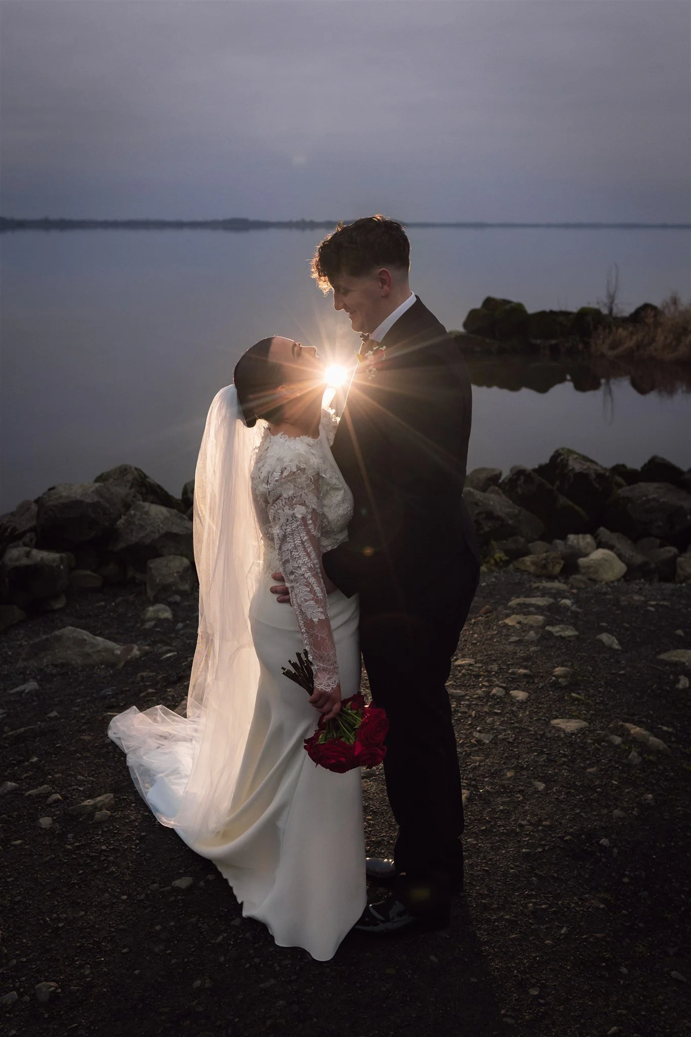 A bride and groom standing close together outdoors near a body of water at sunset, with the bride holding a bouquet of red roses, and the couple smiling warmly at each other.