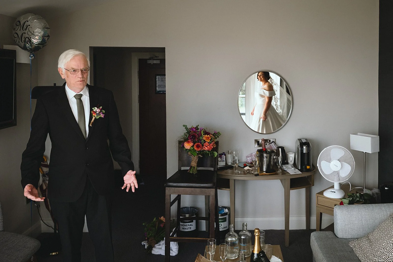 An older man dressed in a black suit with a boutonniere standing in a room decorated for a wedding, with a wedding bride seen in a mirror reflection on the wall.