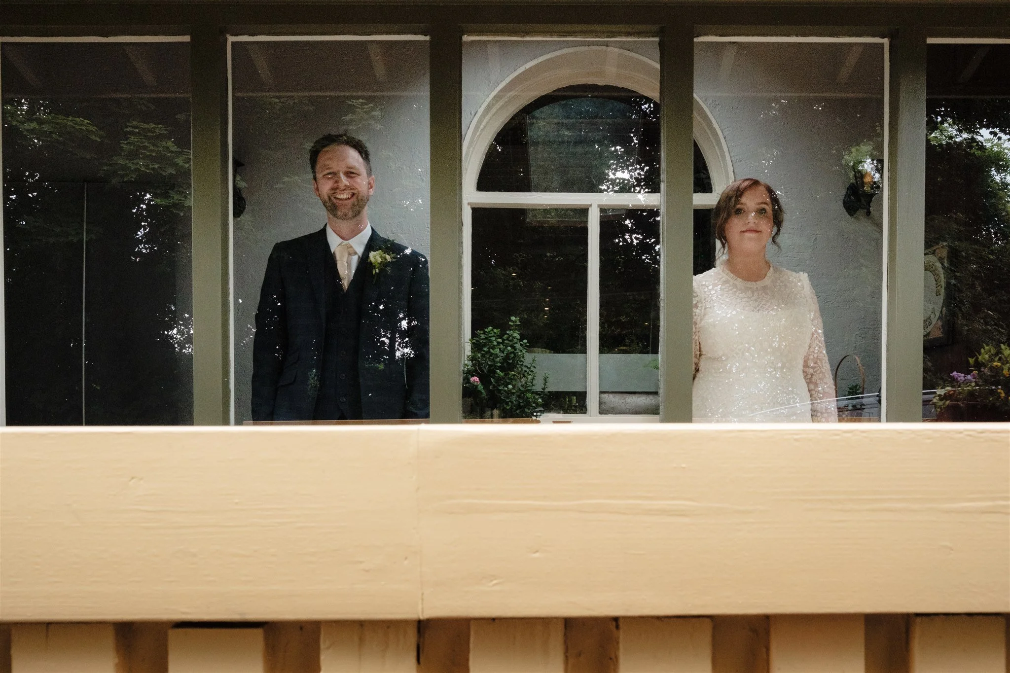 A man in a black suit with a boutonniere and a woman in a white dress are standing behind a window with divided panes. The background features a building with an arched window and greenery outside.