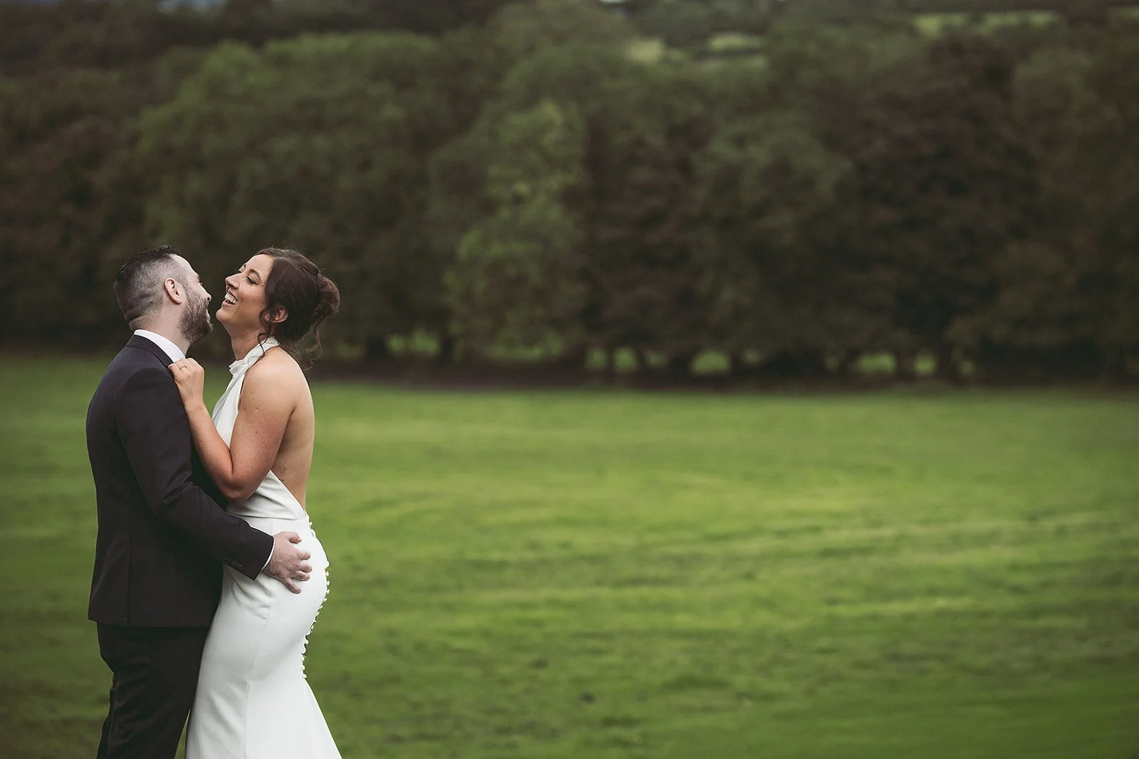 A newlywed couple laughing and embracing outdoors on a lush green field with trees in the background.