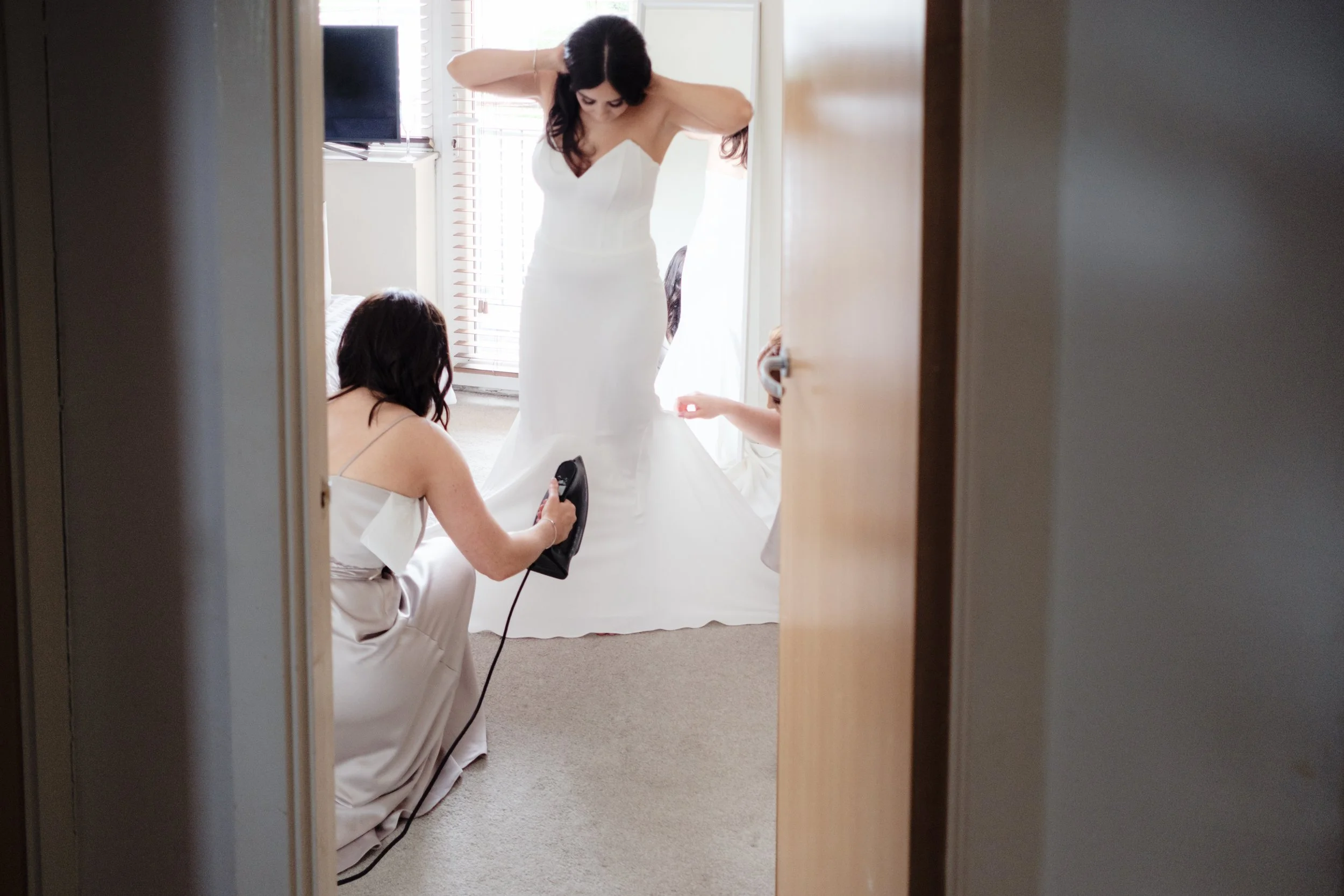 A woman in a wedding dress being assisted by two women as she prepares for her wedding day, seen through a partially open door.