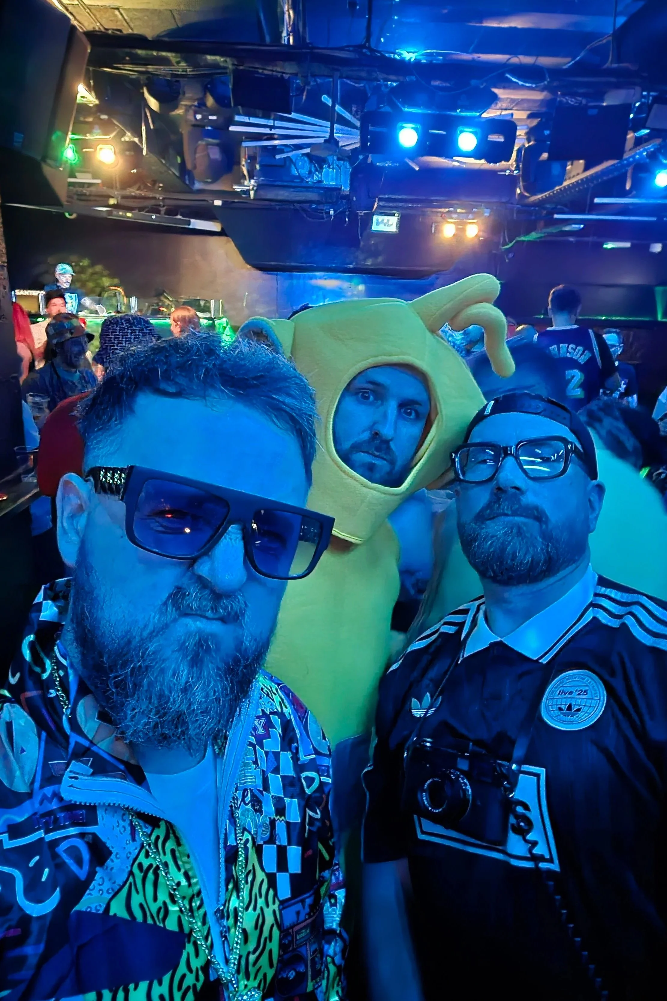 Three men in a club or concert setting, one in a colorful patterned jacket with sunglasses, one in a black mesh shirt with glasses and a camera, and one in a yellow teletubby costume, with a DJ in the background and blue stage lights overhead.
