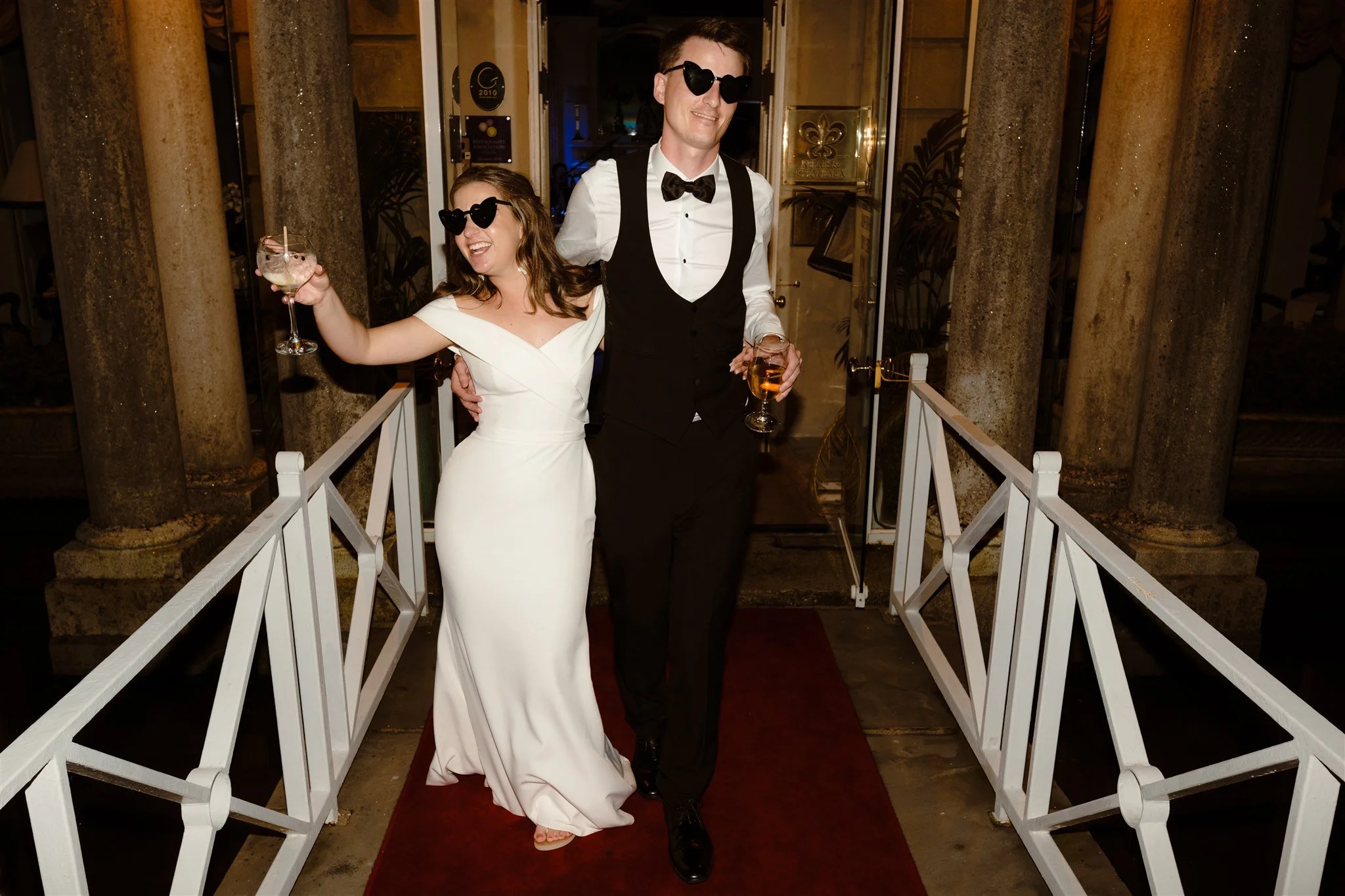 A man and woman dressed in wedding attire, both wearing heart-shaped sunglasses, standing on a red carpet outside a building, holding drinks, smiling, and posing for a photo at night.