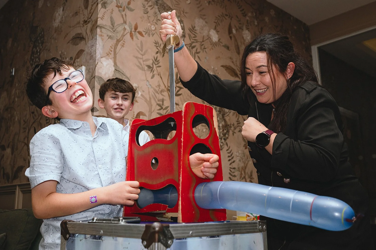 A woman and two boys enjoying a game of Plinko where a plastic tube directs a disk down a board with holes, one boy is smiling with eyes closed and wearing glasses, the woman is holding a hammer, and the second boy is in the background smiling, all a