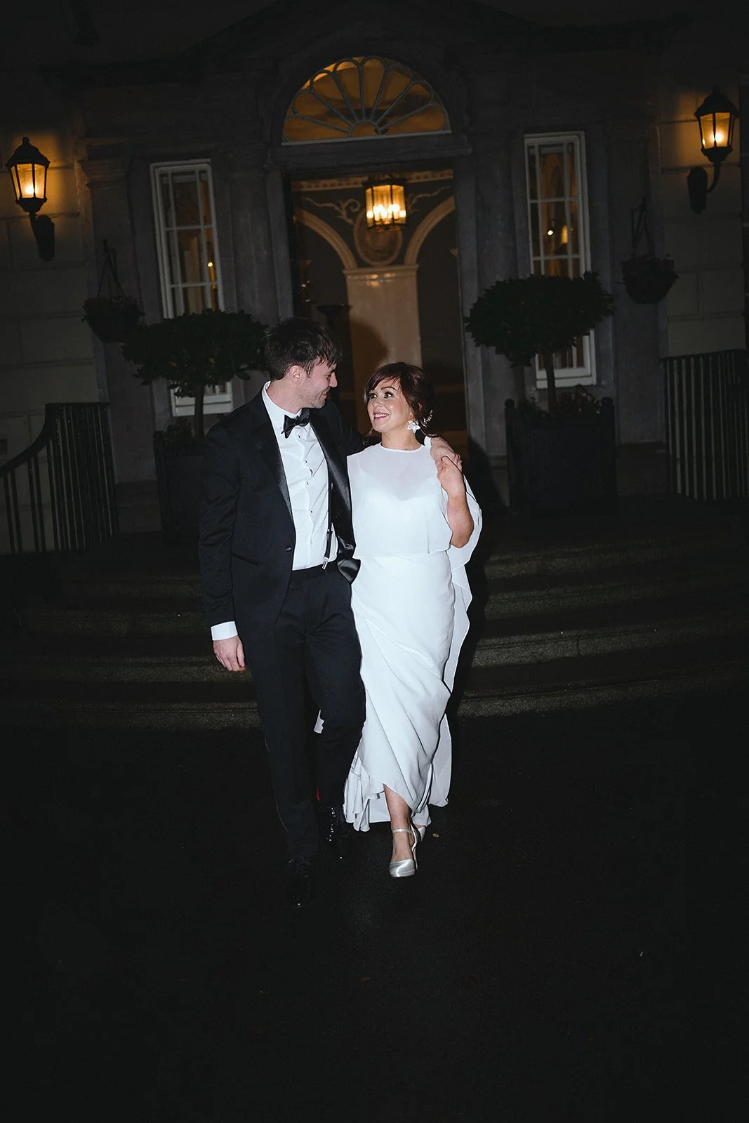 A couple dressed in formal wedding attire walking outside a building at night.