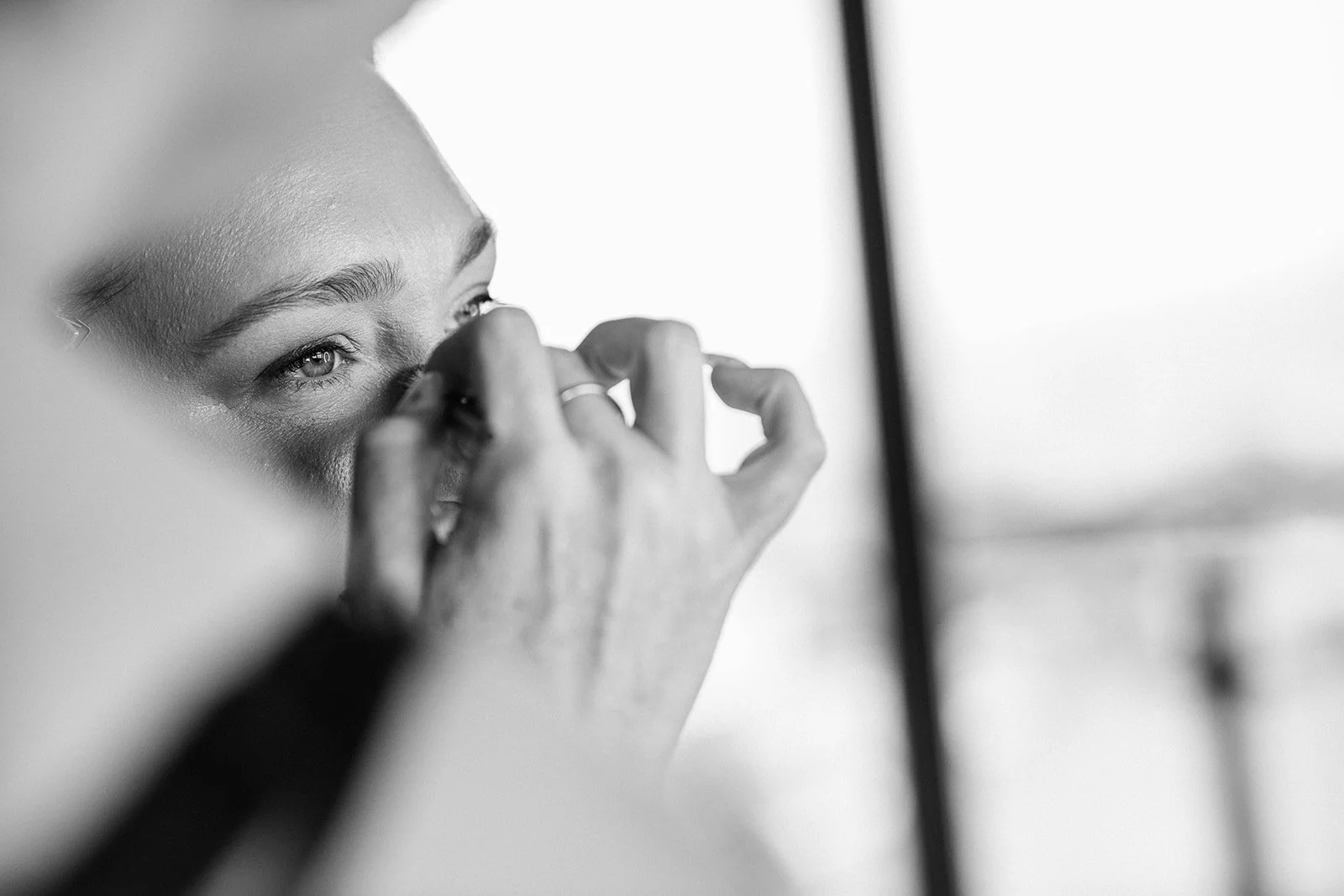 Close-up of a woman applying makeup in front of a mirror, focused on her eye, captured in black and white.