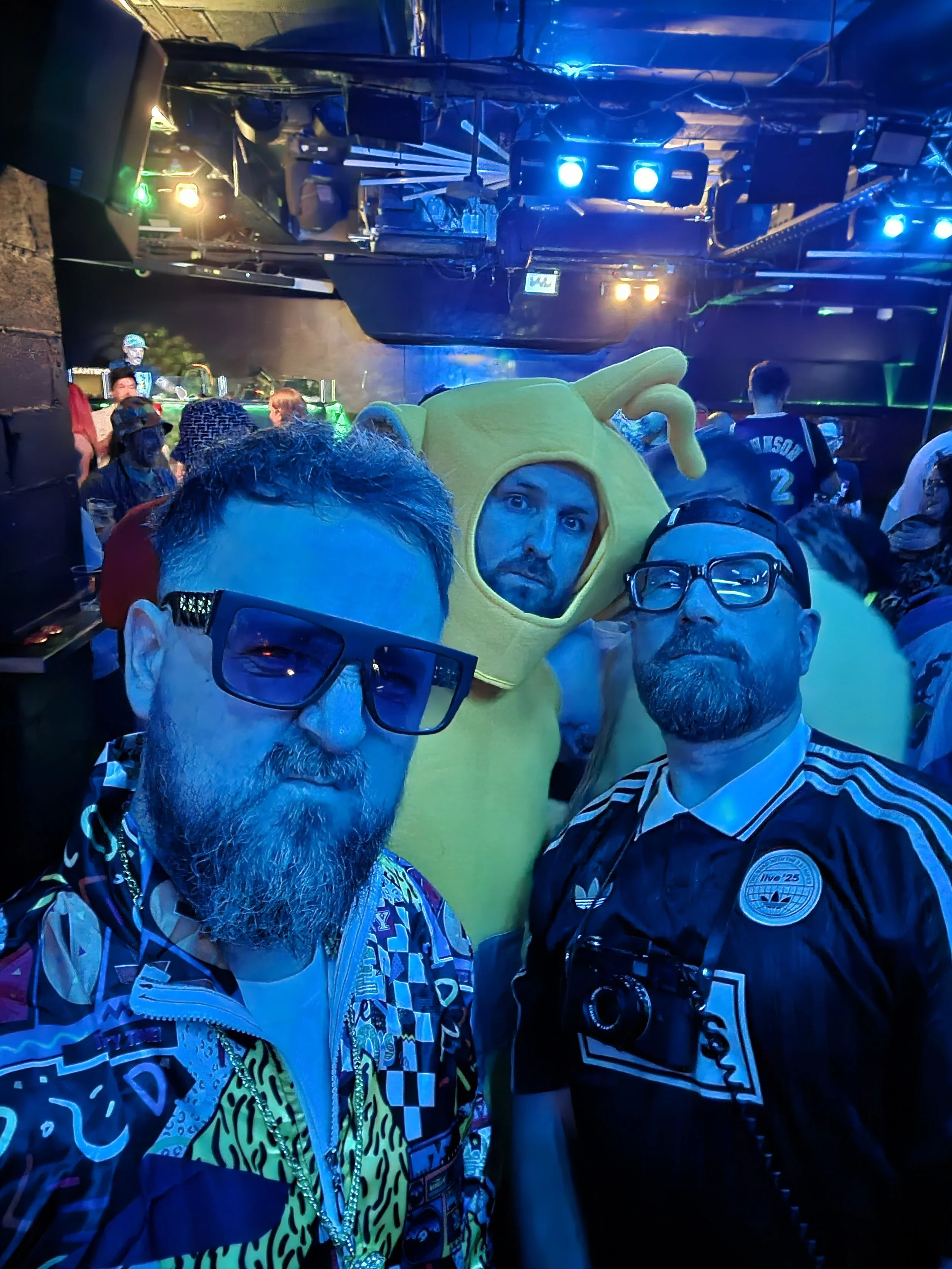 Three men posing for a photo in a crowded nightclub or concert venue with colorful lighting. One man is wearing large sunglasses and a colorful patterned jacket, another is in a black sports jersey with glasses, and the third is dressed in a yellow teletubby costume.