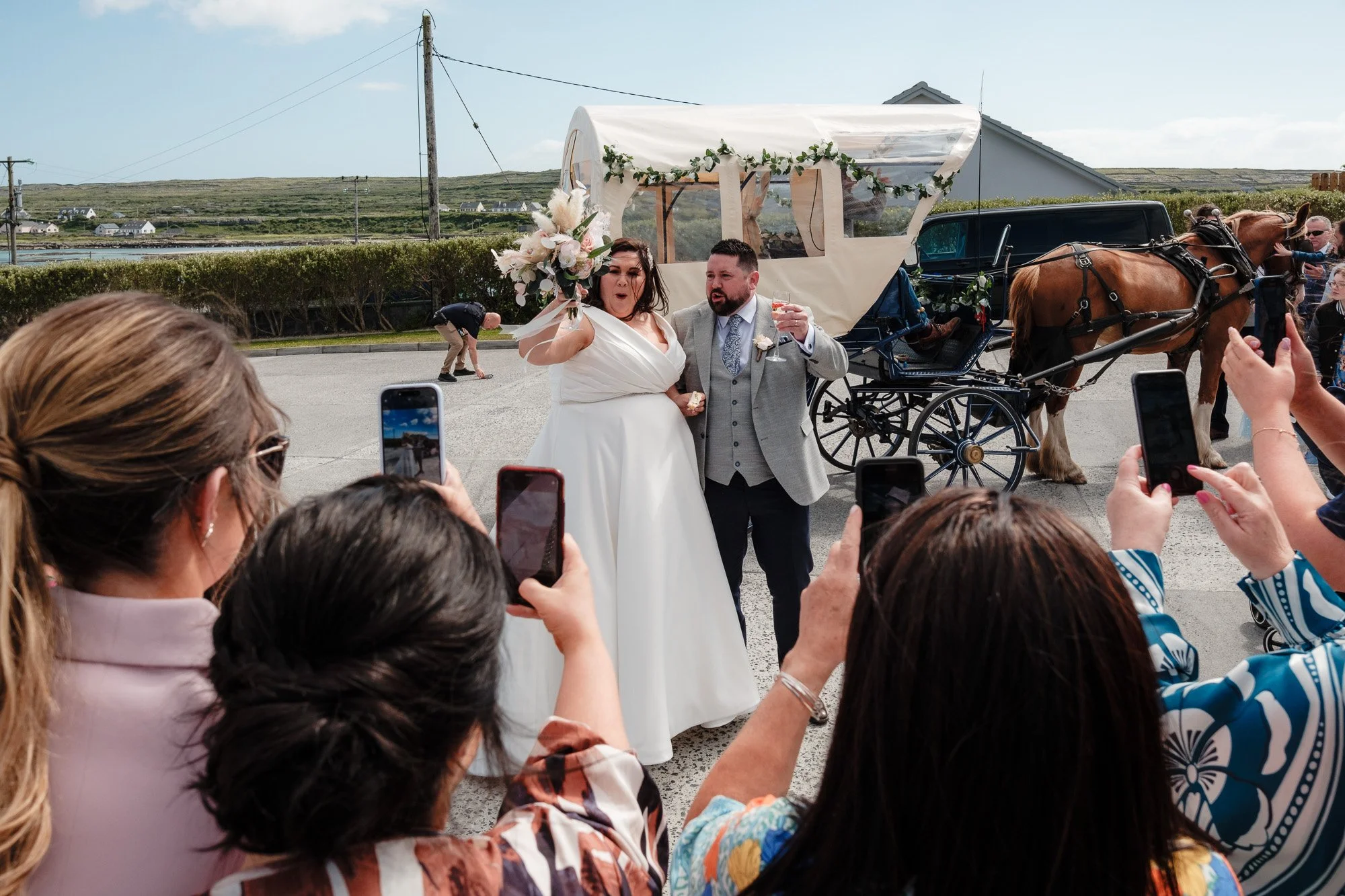 A newlywed couple stands in front of a horse-drawn carriage, surrounded by people taking photos with smartphones. The bride holds a bouquet, and the groom is beside her. The scene is outdoors with a clear sky and a rural backdrop.