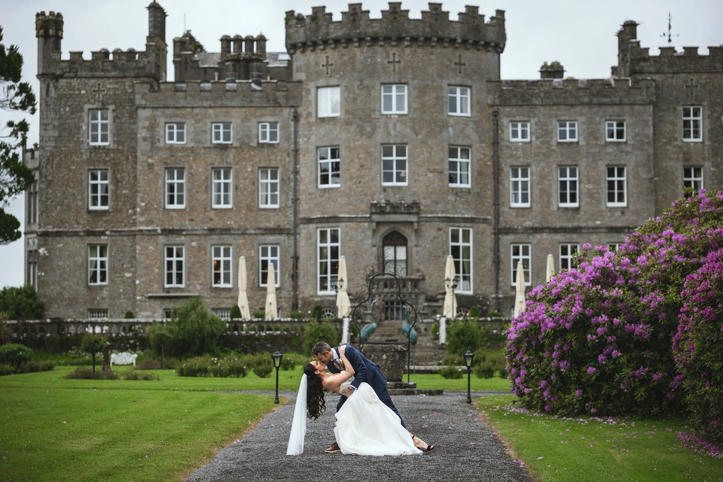 A bride and groom kissing on a gravel path in front of a castle, with greenery and purple flowers Nearby.