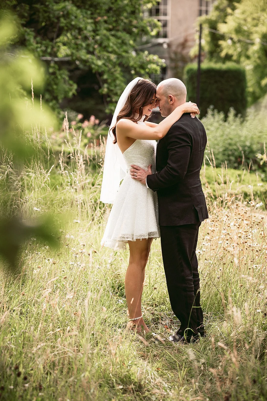 Bride and groom embracing in a grassy outdoor setting, with trees and a building in the background.