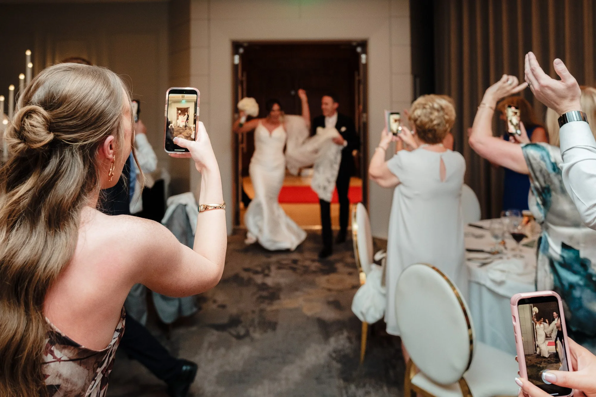 A bride and groom walking out of a building to a room filled with seated women taking pictures of them using their phones, at a wedding reception.