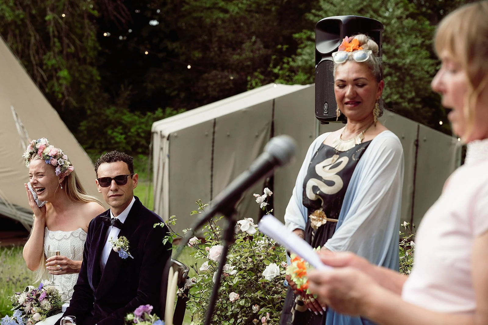 A woman giving a speech at an outdoor wedding ceremony, overlooking a seated bride in a floral crown, groom in a tuxedo, and another woman with sunglasses in front of a wooded backdrop.