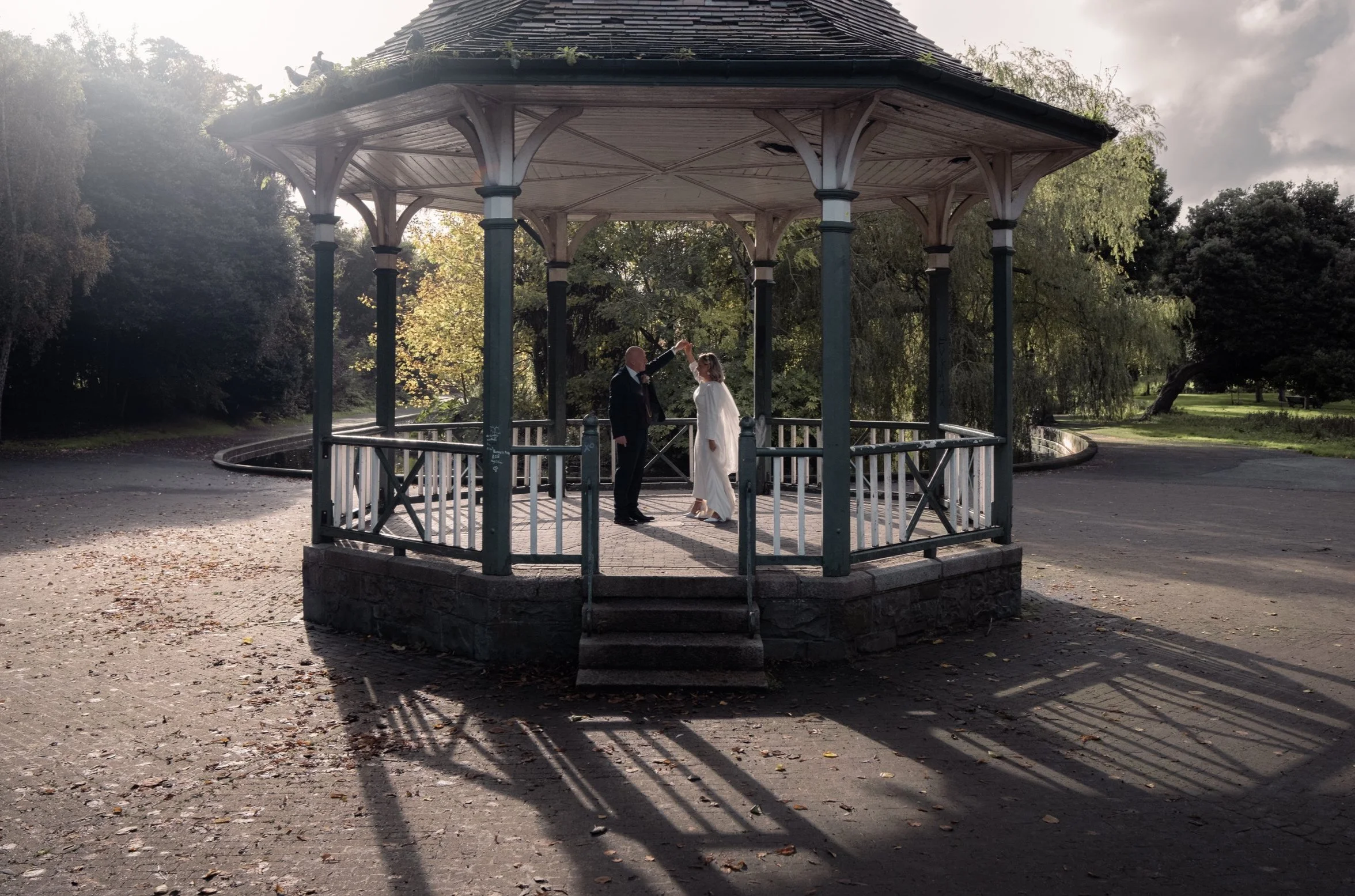 A small, open-air gazebo with ornate support beams, situated in a park surrounded by trees and greenery, with two people dressed formally under it, possibly in a romantic or ceremonial setting.