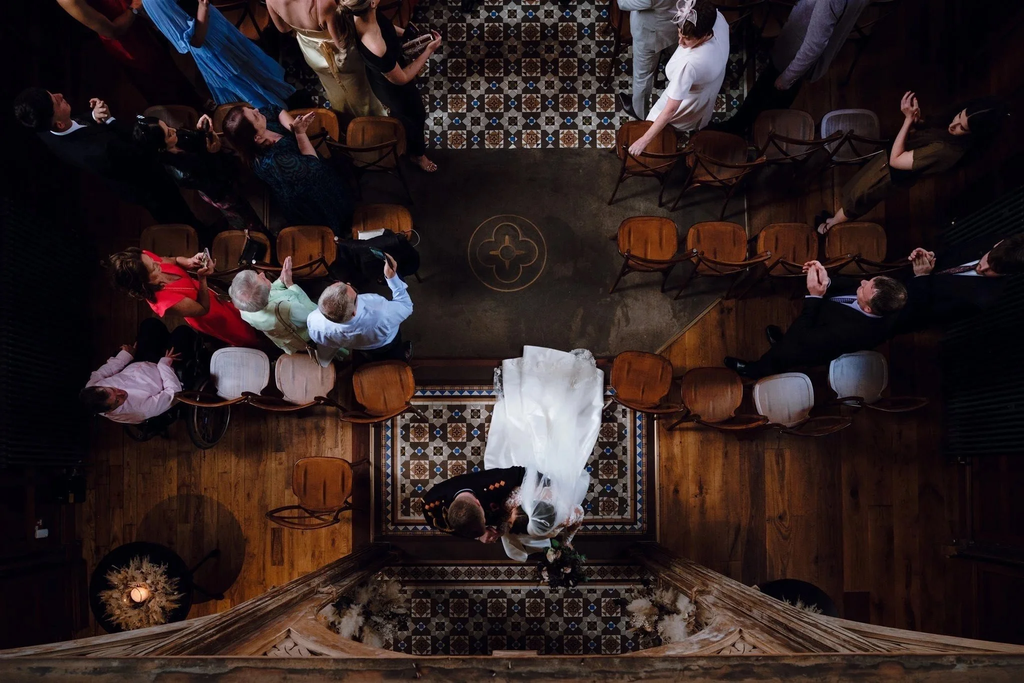 Top-down view of a wedding ceremony showing the bride and groom kissing at the altar, surrounded by seated guests in a decorated indoor space.