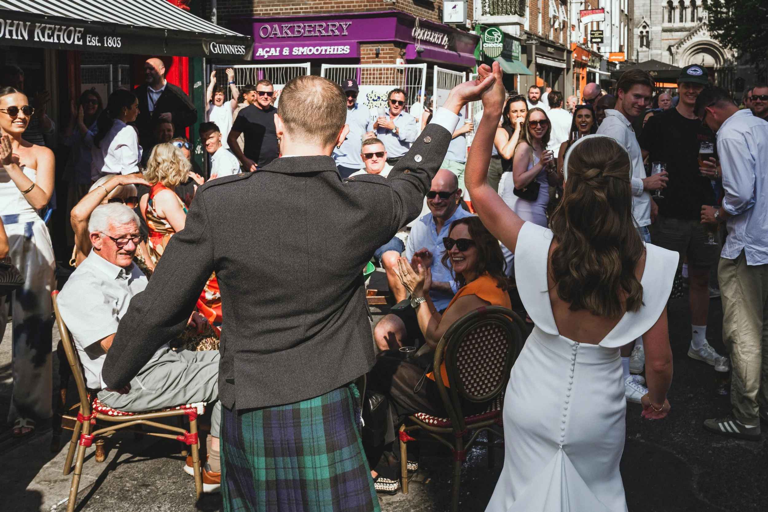 A lively outdoor gathering on a sunny day with many people socializing and enjoying drinks. A person in a suit with a plaid kilt is gesturing with their hand raised, and a woman in a white dress faces them. The scene takes place in a street with vari