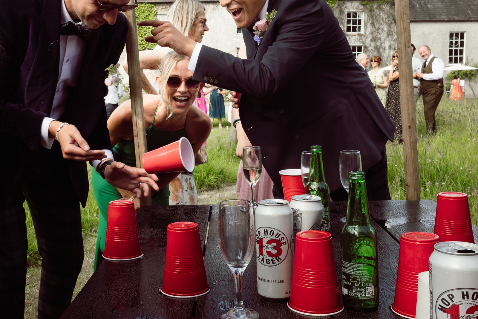 People celebrating at an outdoor party with red cups, beer bottles, and cans on a wooden table, laughing and having fun.