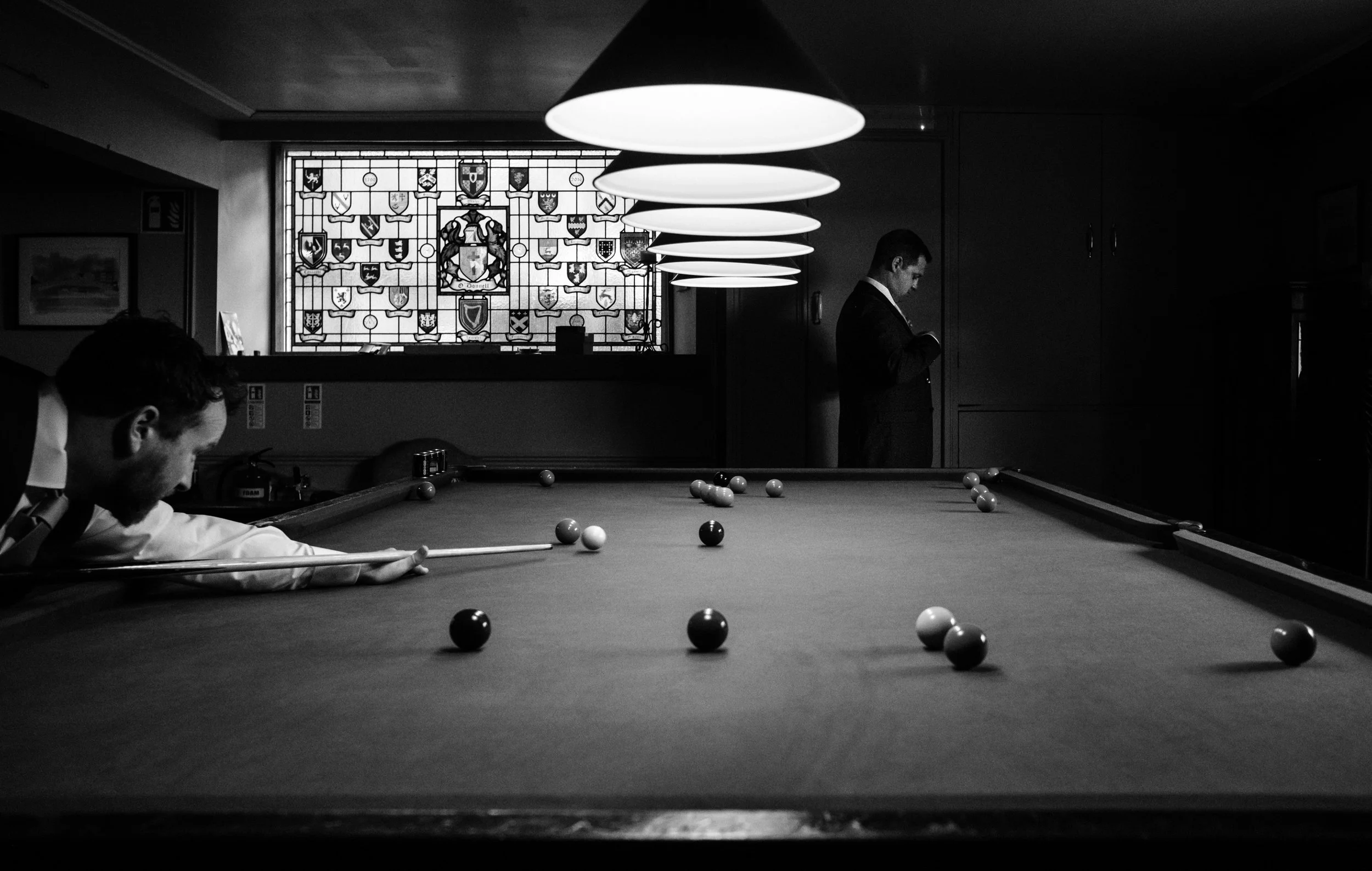 A black-and-white photo of a billiards game in progress, with players aiming at the balls on the pool table, a large stained glass window with coats of arms in the background, and a man standing near the wall